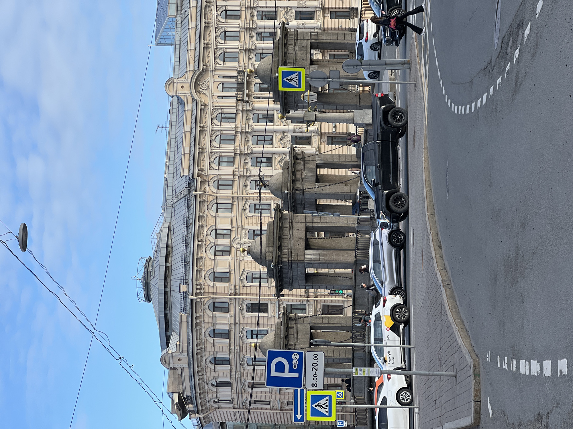 The image shows a street scene in Saint Petersburg, Russia, with a large ornate building dominating the left side. The building features a light-colored facade, rows of arched windows, and decorative architectural elements. Alongside the building, on the right side of the image, there is a line of parked vehicles including taxis and a dark pickup truck. A pedestrian crosswalk sign is visible along with a parking sign marked "8.00-20.00" with "10/15/20" above the parking symbol. A person in dark clothing is walking near the vehicles at the front of the image. A curb separates the sidewalk from the roadway, and there are dashed white lines painted on the road. Overhead, power lines extend across the sky.
