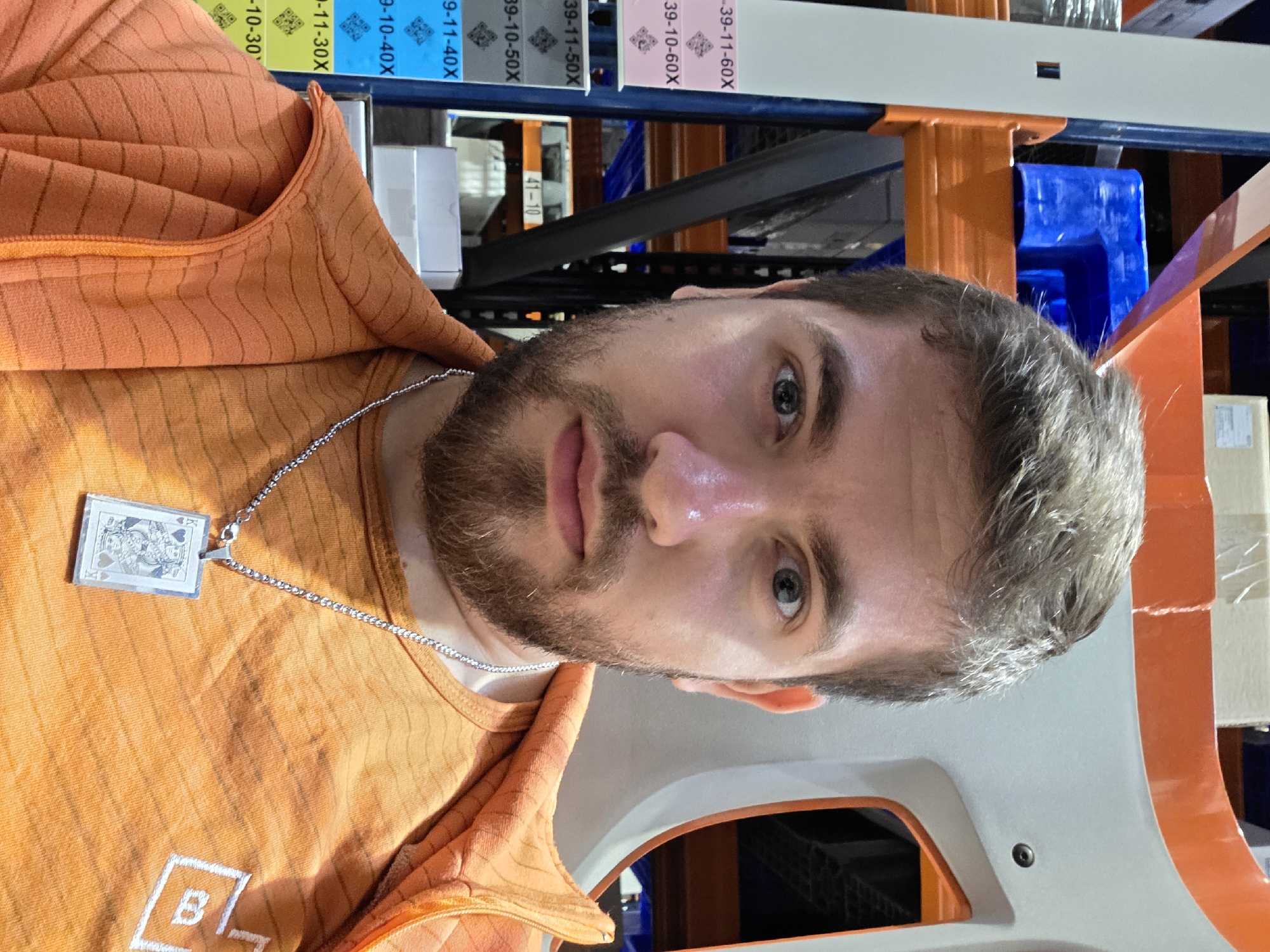 A close-up portrait of a man wearing an orange t-shirt and a silver necklace with a playing card pendant. He has a beard and graying hair. The background appears to be inside a warehouse or storage facility, with shelves and storage racks visible. Labels on the shelves are partially visible, with numbers like "39-11-60X", "39-10-50X", and similar codes. There is no specific indication that the location is Plovdiv, Bulgaria; this information is not discernible from the image.