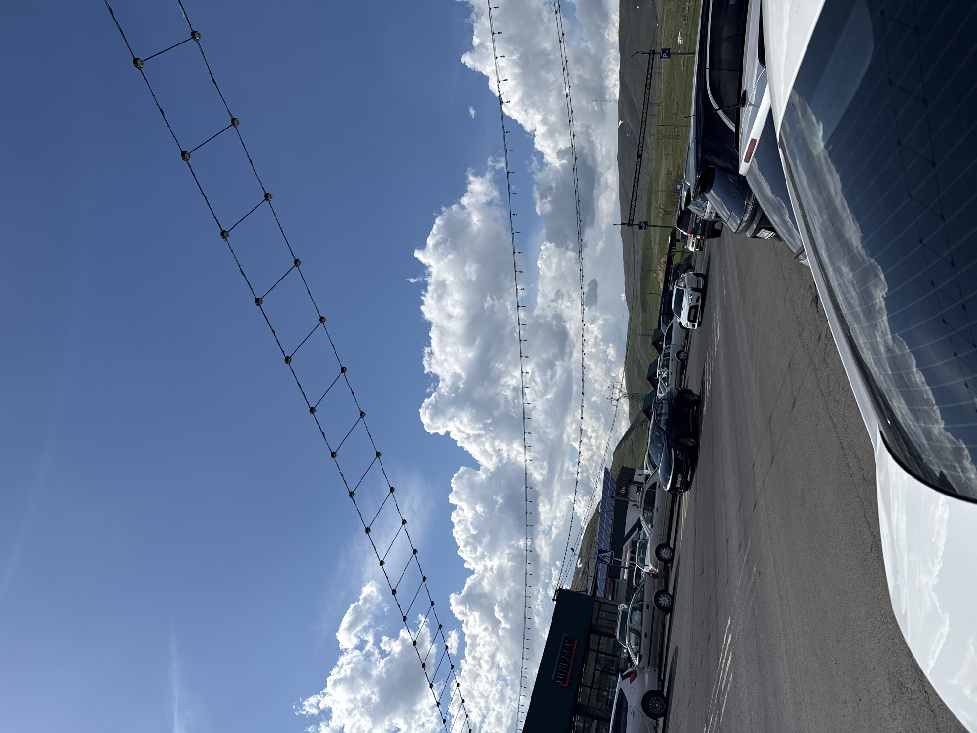 The image shows an outdoor scene in Tsovagyugh, Armenia. A parking lot with multiple parked cars is visible along a paved road. Behind the parking lot, a grassy hillside stretches towards the horizon. Overhead, the sky is mostly blue with fluffy, white cumulus clouds. Two types of overhead lighting structures are visible. One is a simple string of lights. The second is a grid of cables with evenly spaced dark connectors. The lighting structures are suspended above the parking lot. A building with the name "UNIPACK" can be seen. The image is oriented sideways.