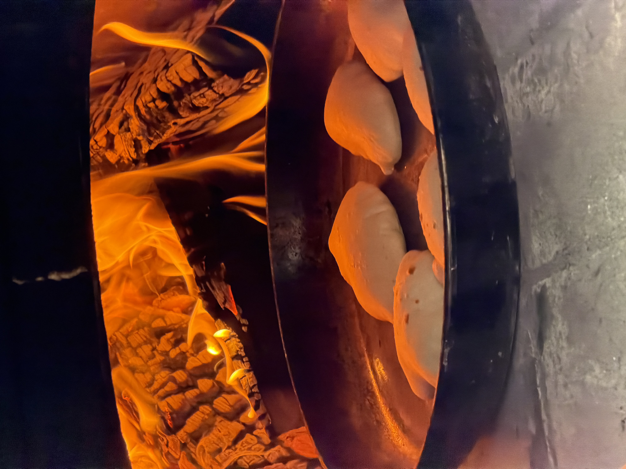 The image shows a close-up view of dough balls being cooked in a rustic oven, likely a wood-fired oven or a forge. The scene is dominated by the warm glow of a fire on the left side, with visible logs and vibrant orange flames licking upwards. The heat from the fire is casting a warm light on the dough balls arranged in a metal pan or on a flat surface to the right.
The dough balls are pale and appear to be in the process of baking or cooking. Some show slight browning or variations in texture, hinting at the cooking process. The pan or surface holding the dough is dark and metallic, with a curved edge visible. To the far right, a textured, grey surface is visible, possibly the interior wall of the oven or an adjacent structure.
The lighting suggests it could be evening or indoors, where artificial light or the fire is the primary source. The overall mood is cozy and industrial, evoking a sense of traditional cooking or craft. There is no visible text in the image.