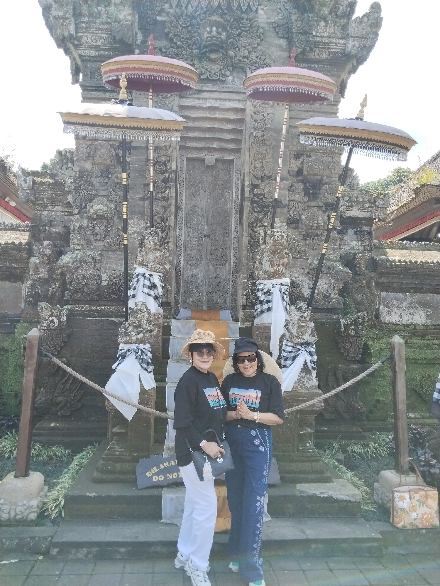 In Bangli, Indonesia, two women stand on a stone pathway in front of a large, intricately carved stone temple structure. The temple has a prominent gateway with two large, ornate umbrellas displayed on either side. The umbrellas are multi-tiered with red and gold accents and are supported by dark poles. The temple's facade is adorned with detailed carvings and flanked by stone statues wrapped in traditional black and white checkered fabric. White fabric is also draped around the bases of the statues and pillars.
The woman on the left wears a straw hat, sunglasses, a black long-sleeved shirt with a colorful graphic, white pants, and athletic shoes. She carries a black handbag. The woman on the right wears a dark baseball cap, sunglasses, a black long-sleeved shirt with a graphic, and blue pants with embroidered floral patterns. She wears green-accented shoes and stands with her hands pressed together in front of her chest.
In the foreground, a sign reads "DILARANG DO NOT". Green foliage grows around the base of the temple, and a rope barrier runs across the scene. A patterned tote bag rests on the ground to the right of the women. The sky is bright and appears to be a clear day.