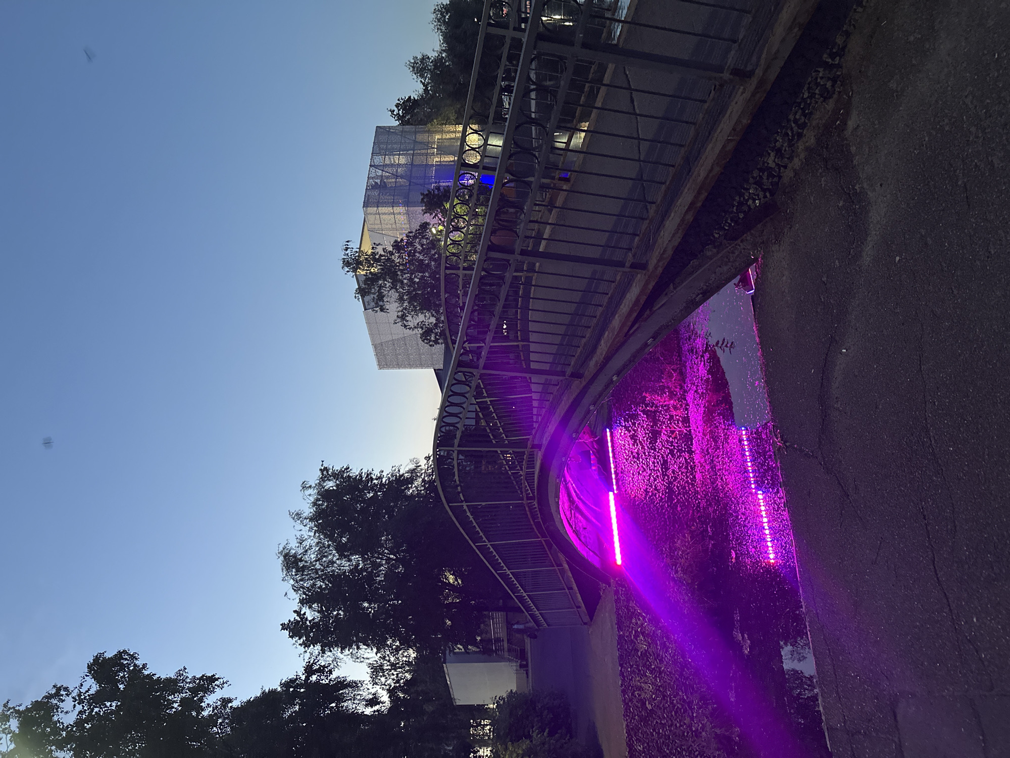 The image captures an evening scene in Kazan, Russia, featuring a bridge over a body of water illuminated by vibrant purple lights. The perspective is from alongside a dark asphalt path, looking towards and under the bridge.
In the foreground and midground, a stream or canal runs parallel to the path, its water and the adjacent vegetation intensely lit by a series of hidden purple LED lights, casting a strong glow and reflections. A metal bridge or elevated walkway with an ornate, dark railing curves across the frame, leading towards the upper right. Below the bridge, the structural elements are visible, and some natural debris like small rocks are present.
Beyond the bridge and partially obscured by trees, a modern building with a distinctive mesh-like or glass facade is visible, distinguished by internal blue lighting. The sky above is a clear, light blue, suggesting either dusk or dawn, with enough natural light remaining to outline the silhouetted treetops. The overall impression is one of a public space or park designed with contemporary architectural lighting to create a striking nocturnal ambiance. There is no visible text or human activity.