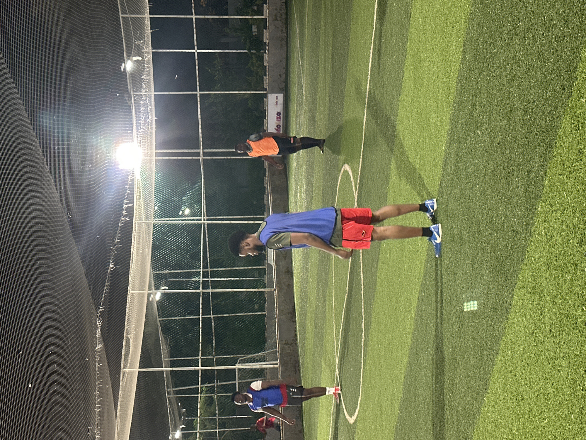 In Abuja, Nigeria, an outdoor, artificial turf soccer field is illuminated by bright overhead lights. Three men are visible on the field, engaged in what appears to be a soccer practice. The man in the foreground, facing away from the camera, wears a green t-shirt and red shorts. To his left, another man in a blue bib and grey shorts stands facing him. Further in the background, a third man in an orange bib and black shorts is positioned on the field. The field is marked with white lines, including a center circle. Fencing surrounds the field, with a net overhead, suggesting it's an enclosed sports facility. The lighting suggests the game is taking place at night.