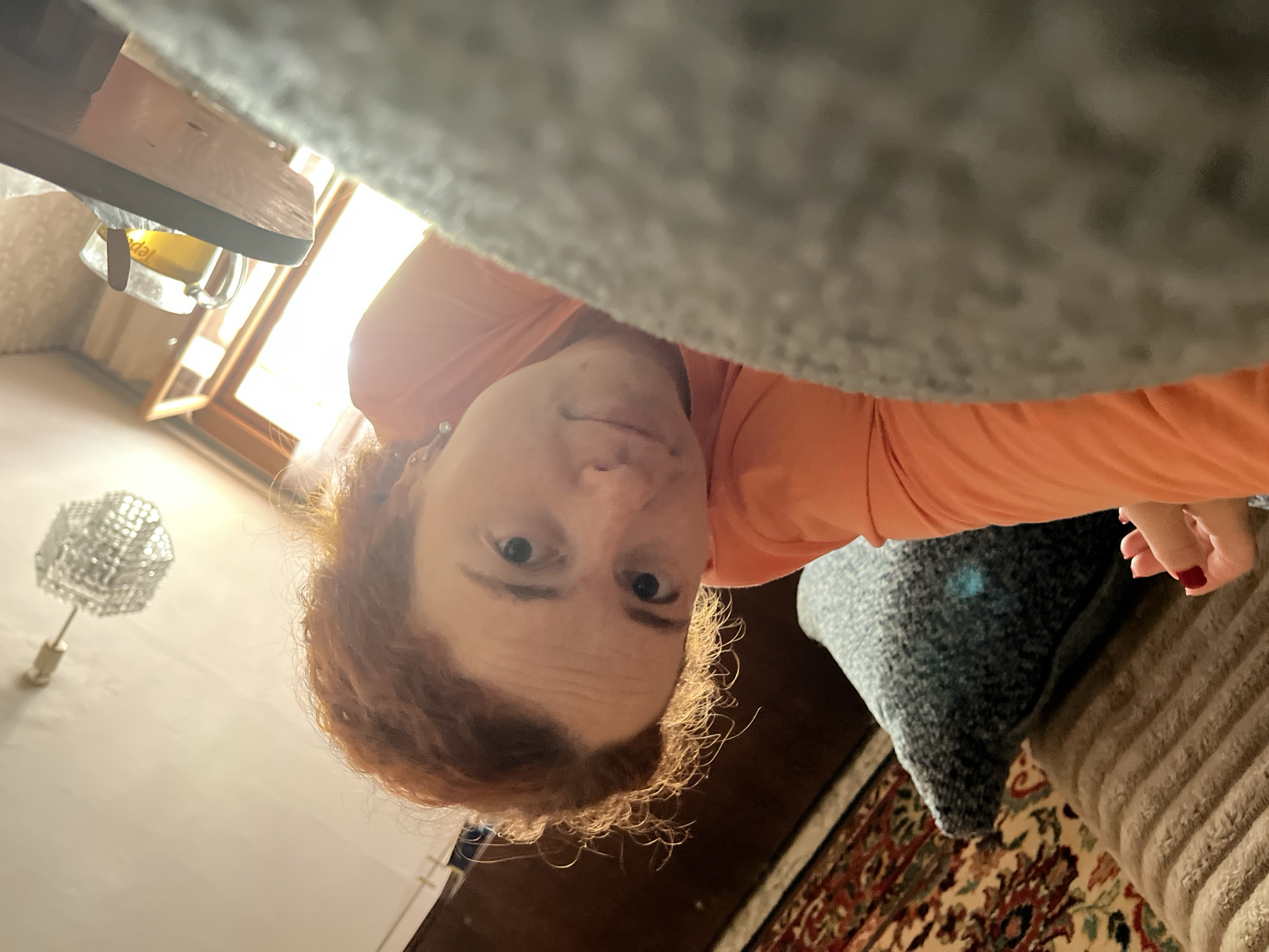 The image is an upside-down selfie of a fair-skinned person with reddish-brown hair. They are wearing an orange shirt. The background includes a partially visible window with sunlight streaming through, a table with a glass mug on it, a wall lamp, and a section of a patterned rug. The lower right shows parts of couch cushions. The location is Chaikovsky, Russia.