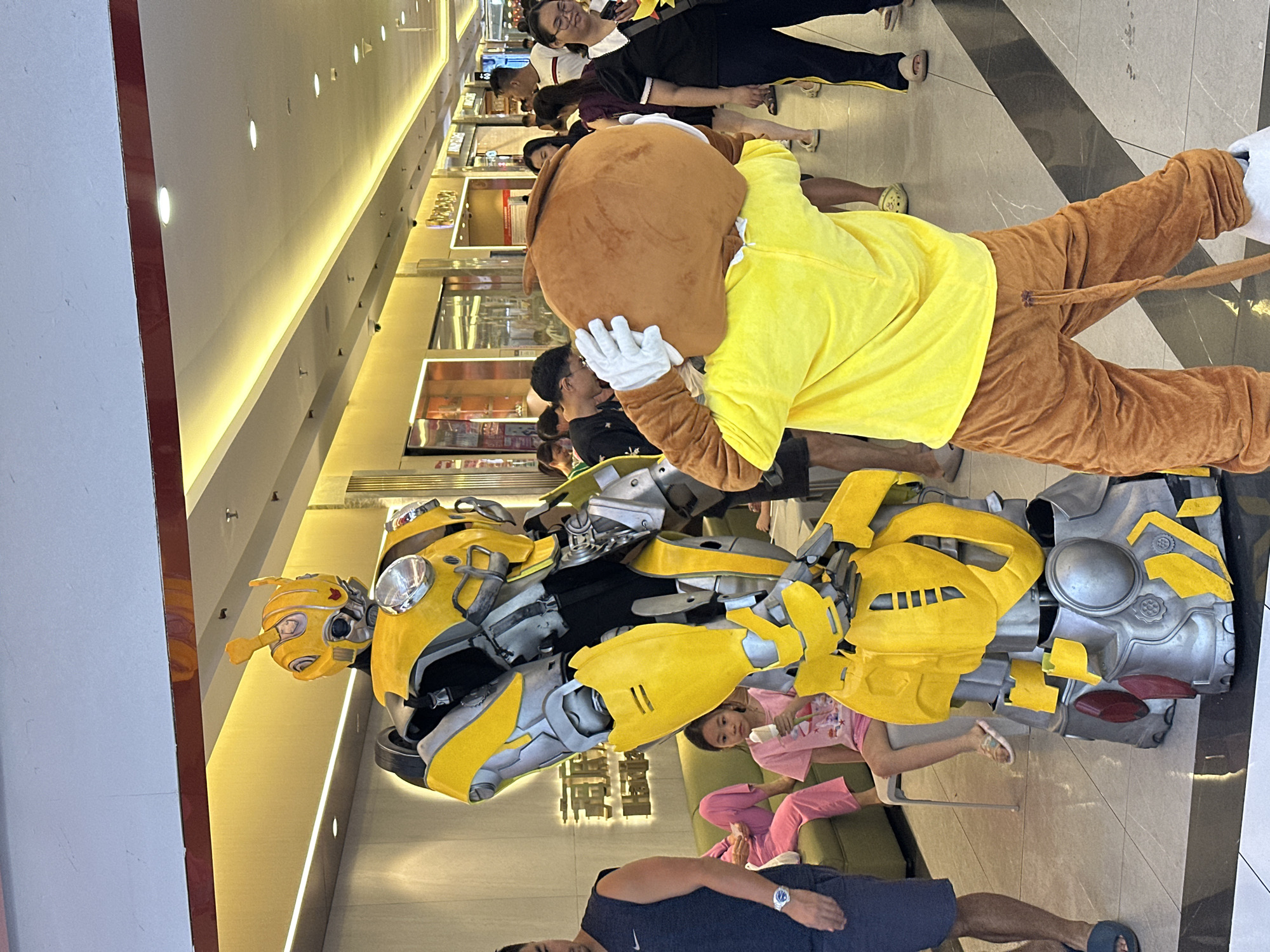 The image depicts an indoor scene, likely in a mall, in Thủ Đức, Vietnam. A person in a Bumblebee costume stands prominently in the foreground, positioned next to a person in a Lion costume. Several people are gathered around the costumes, some appearing to observe or pose for photographs. A child in pink clothing is sitting on a green chair in the vicinity of the costumes. The location appears to be a well-lit, modern commercial space, possibly inside a mall, in Thủ Đức, Vietnam. The floor is tiled in a pattern of light and dark colors. Other shops and people can be seen further in the background.