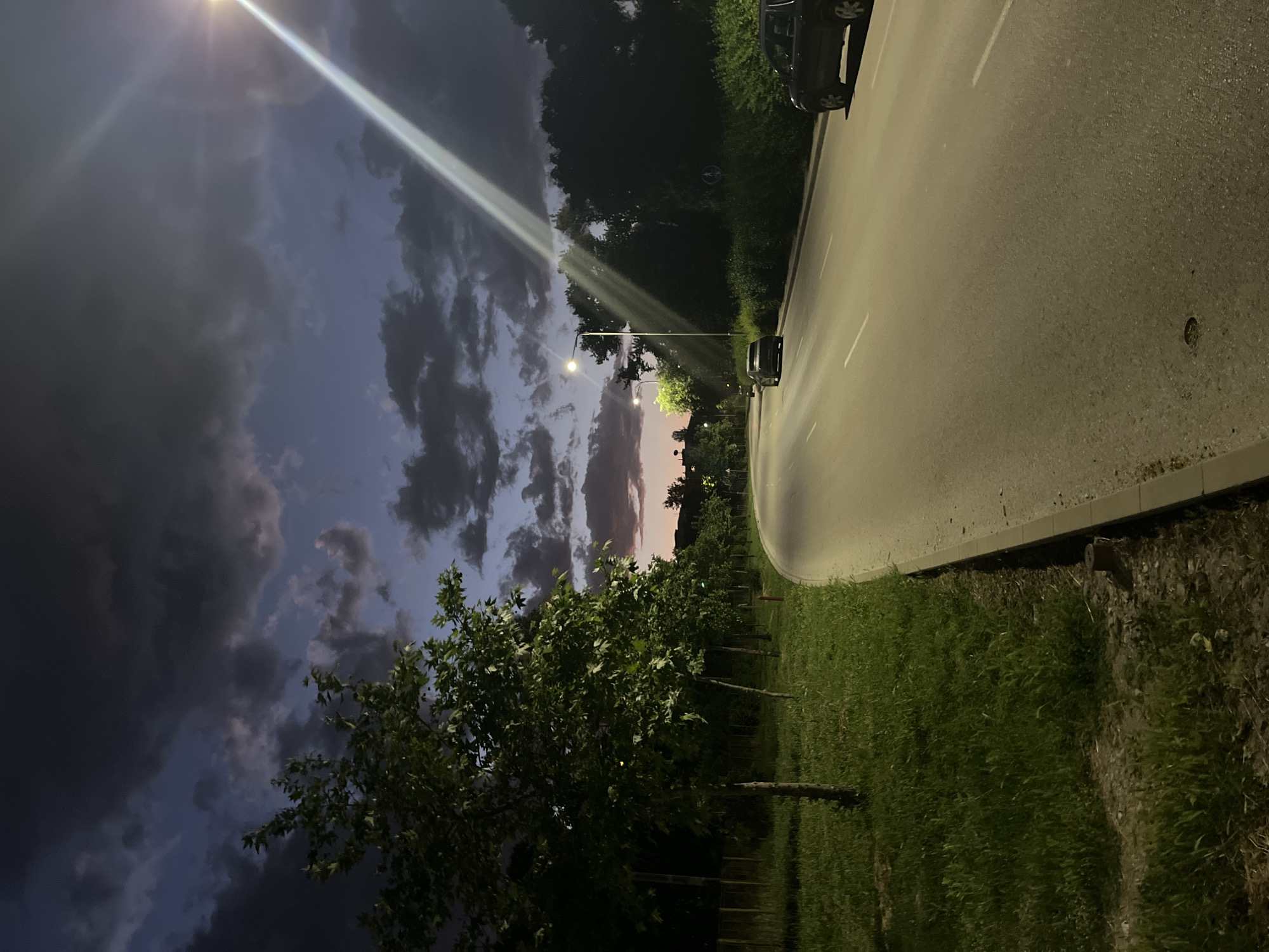 The image depicts a nighttime scene in an urban environment. A street curves into the distance, illuminated by streetlights. A car is parked on the side of the road. Trees line the opposite side of the street. The sky is filled with dark clouds, and a ray of light shines through them. The location is Unknown, Unknown.