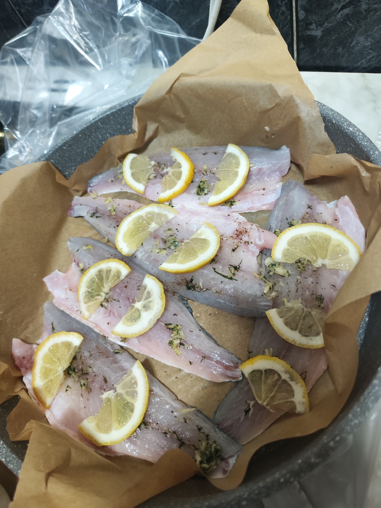 The image depicts raw fish fillets arranged in a pan lined with parchment paper, topped with lemon slices, herbs, and possibly garlic. The pan appears to be gray, and in the background, there's a clear plastic bag and a glimpse of what might be a countertop or backsplash. There is no indication of the location of the city of Burgas, Bulgaria.