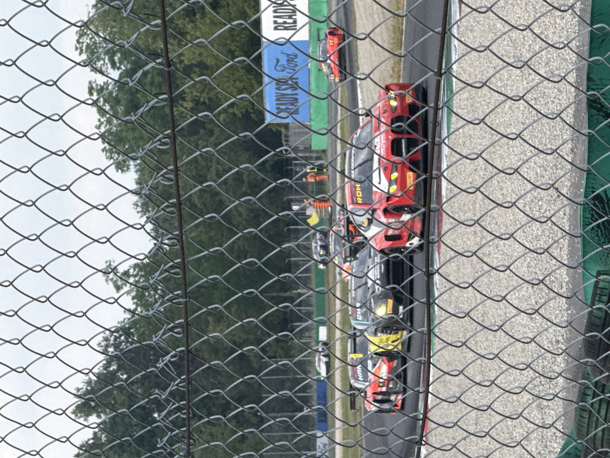 The image, taken through a chain-link fence, shows a motor racing event at the Monza Circuit in Monza, Italy. Several race cars, primarily red and black, are visible on the track. Race officials wearing orange vests are present near the track. A "READY. SET. Ford" banner is also visible. The background includes trees and a grey sky.