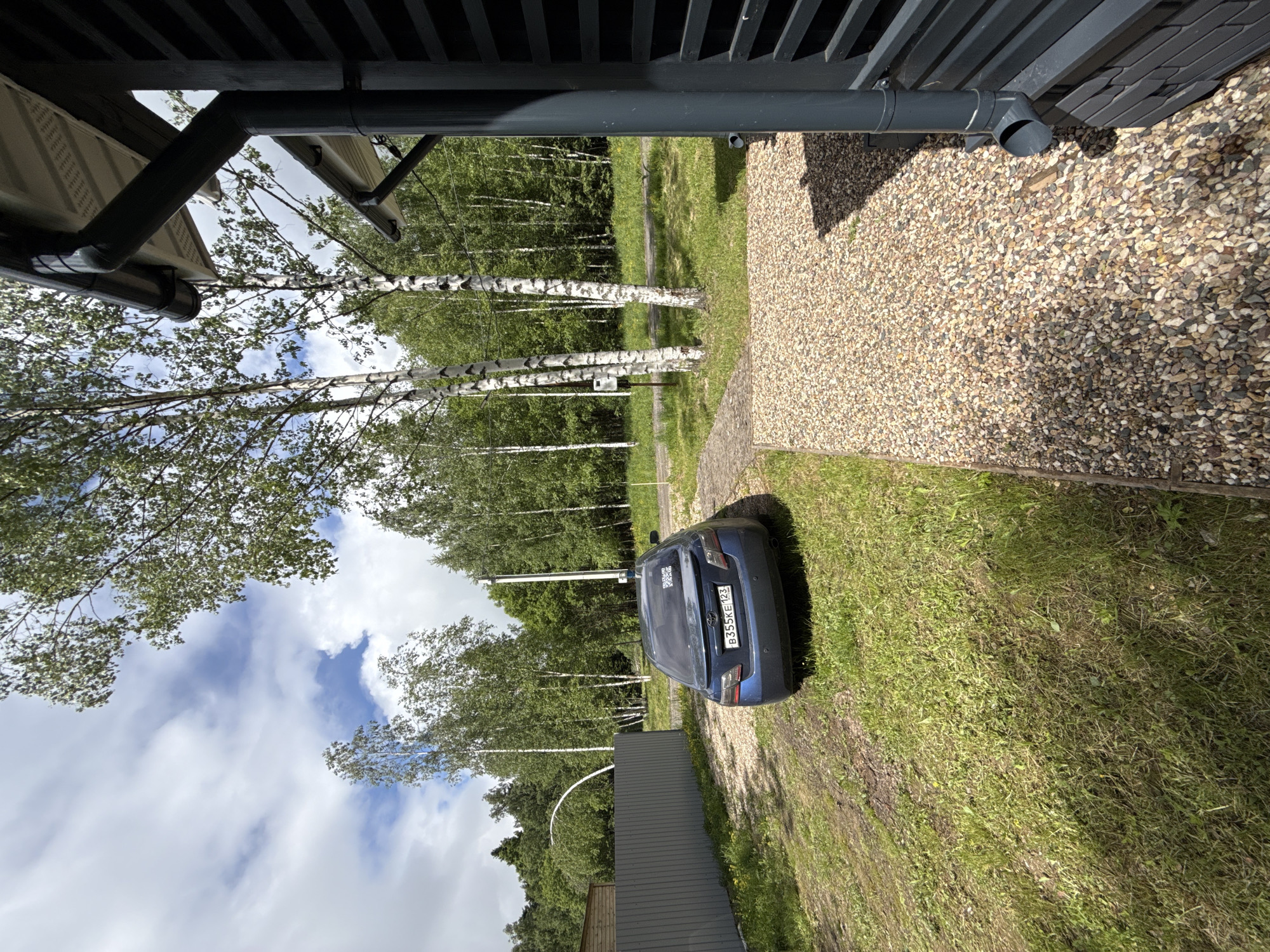 The image depicts a scene at Buzhaninovo, Russia. A blue car, with the license plate B355KE123, is parked on a grassy area beside a gravel driveway. The driveway leads towards a building or structure partially visible in the upper right corner. In the background, birch trees line the area. The sky is partly cloudy. An overhanging structure with gutters is in the top portion of the image, suggesting it's taken from underneath a covered area. A grey fence is on the left side of the car.