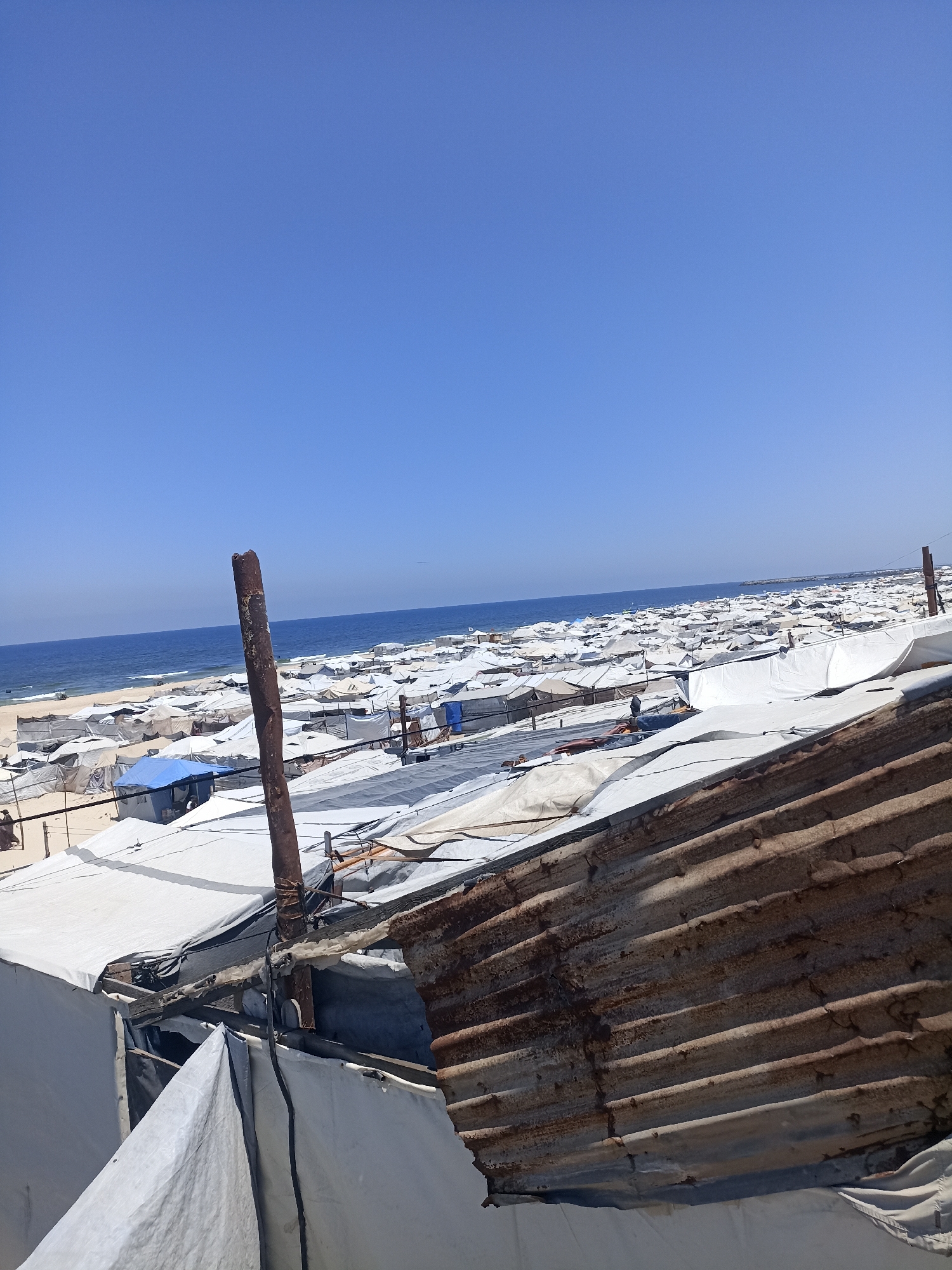 The image depicts a large refugee camp situated near a coastline under a clear blue sky. Numerous white tents, likely makeshift shelters, are densely packed together, stretching as far as the eye can see towards the sea. A wide expanse of calm blue water is visible beyond the camp, meeting the horizon. The sand of the beach is visible between the tents and leading down to the water.
In the foreground, a rusted, corrugated metal structure, possibly a damaged roof or wall, partially obscures the view of the tents. A dark, weathered metal pole stands prominently in the center of the frame, also appearing to be part of the temporary structures. Some tents are made of white tarp material, while others appear to be constructed with different fabrics, including a blue tent visible in the middle ground. The overall impression is one of a crowded and precarious living situation.
The scene suggests a bright, sunny day with no visible clouds. The location is identified as Gaza, though the specific site within Gaza is unknown. No specific activity or event is clearly discernible, but the sheer number of tents indicates a significant population of displaced people. The image conveys a sense of scale and the challenging conditions faced by those living in the camp. There is no visible text in the image.