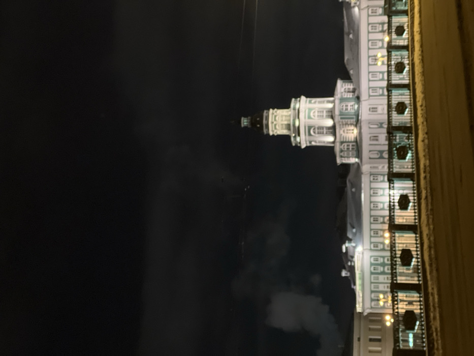 A tower of a building, illuminated at night, stands against a dark sky. The tower is white and green, with a series of windows and architectural details. To the right of the tower is the body of the building, illuminated by lights. The building appears to be beside a sidewalk, which is along the right edge. The dark sky makes up the majority of the background.