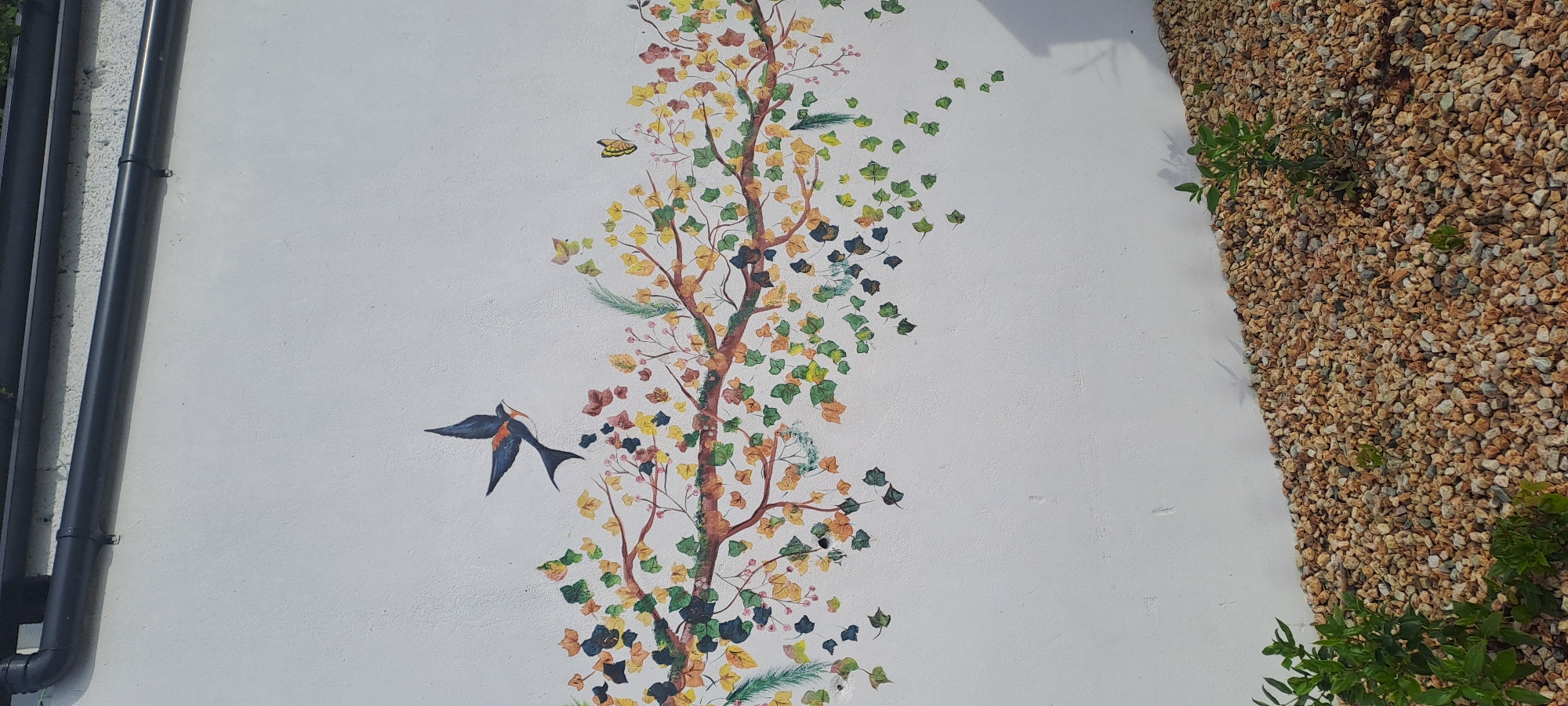 The image shows a white wall with a mural of a tree branch with falling leaves. A bird is depicted in flight above the branch, and a butterfly is visible to the right. Below the wall, there is a ground covered in small, light-colored pebbles. The scene appears to be outdoors, with natural light casting shadows. There are no people or identifiable structures visible in the immediate vicinity that would indicate a specific location within Castlebar, Ireland. The mural is the primary subject, rendered in a stylized, artistic manner.