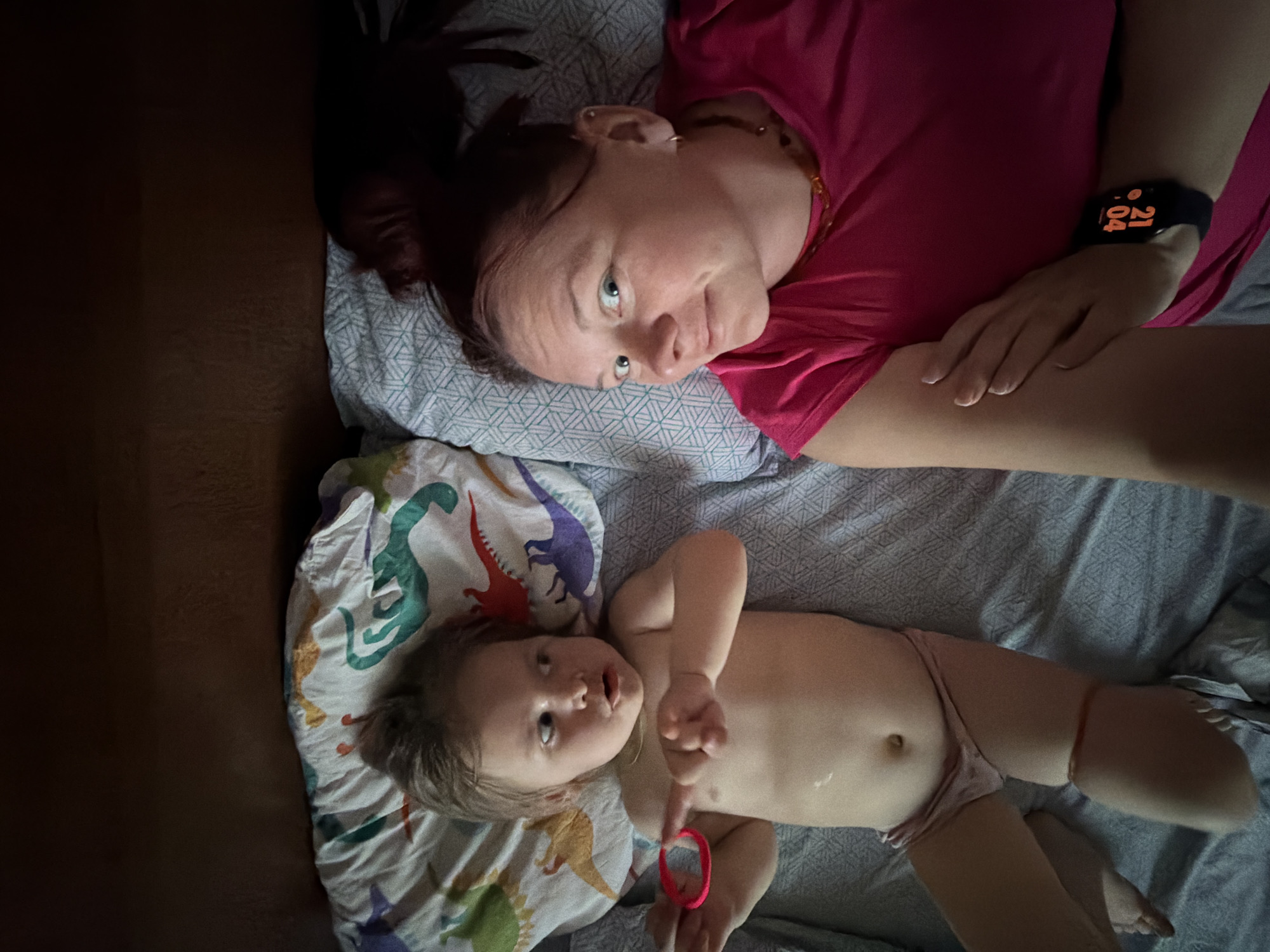 A medium shot depicts a woman and a baby lying on a bed. The woman, wearing a pink shirt and a black smartwatch displaying "21:04", is positioned at the top of the frame, looking towards the camera. The baby, wearing light-colored underwear and holding a red hair tie, is lying on a dinosaur-patterned pillow at the bottom of the frame, also looking towards the camera. The bed has a grey patterned sheet. The left side of the frame is dark. It is impossible to confirm if the location is in the city of Chaikovsky, Russia from the visual elements.