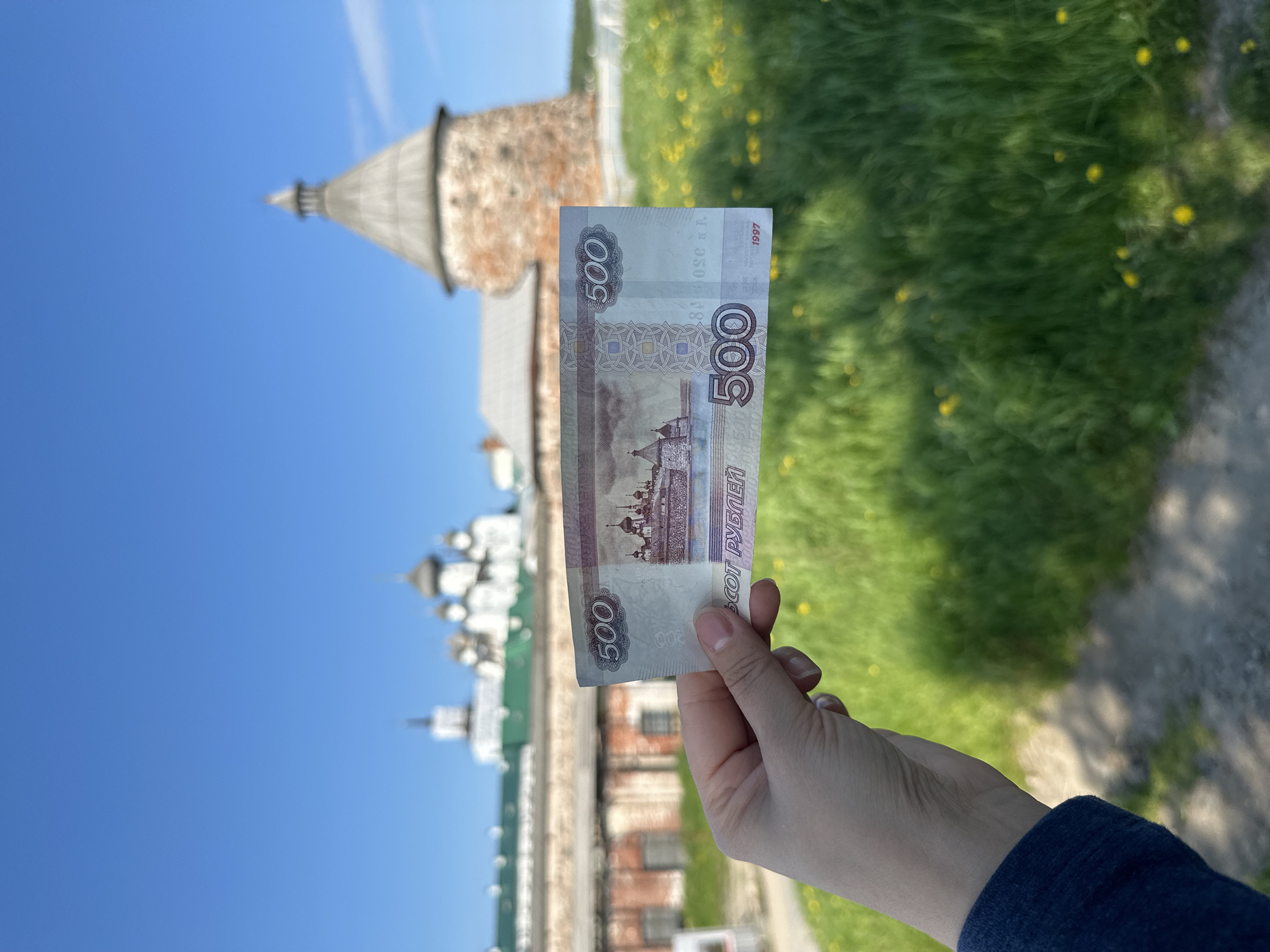 A hand holds a 500-ruble banknote in front of a blurred background. The background features a portion of the Solovetskiy Monastery complex in Solovetskiy, Russia. The monastery includes several whitewashed buildings with domed roofs and a round, stone tower with a conical roof. Green grass with scattered yellow flowers covers the ground in the foreground and midground. The sky is clear and blue.