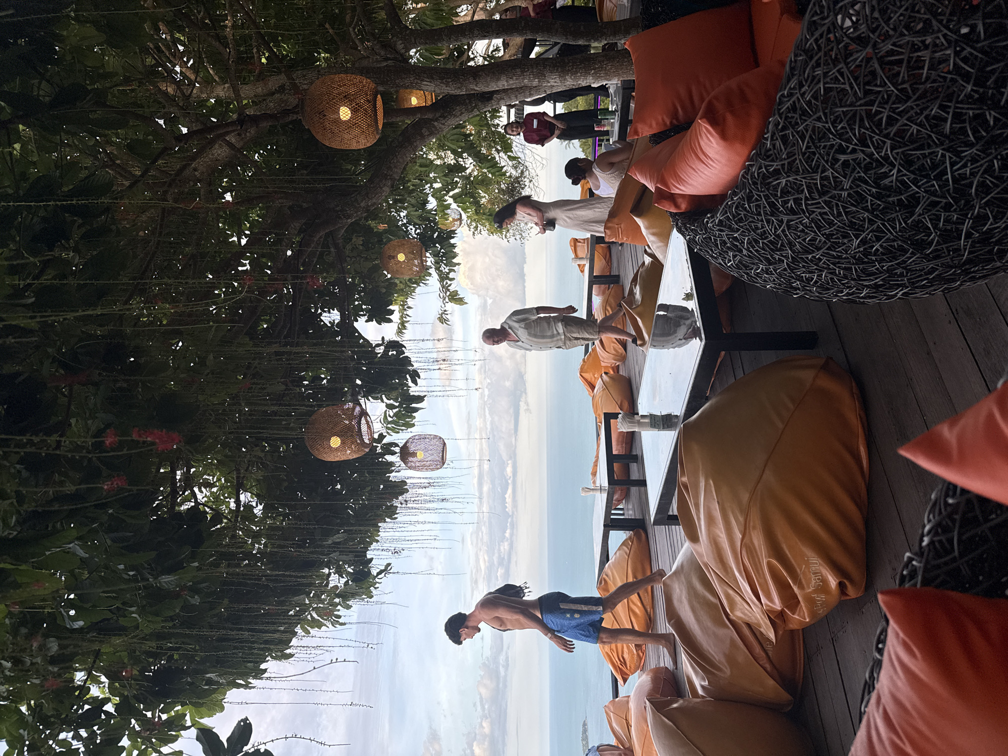 The image depicts an outdoor seating area with a scenic view of the ocean and a cloudy sky. The seating arrangement consists of numerous large, orange beanbag chairs and dark wicker furniture with orange cushions. Several low tables are placed on a wooden deck. In the background, people are walking and standing around the seating area. A man in blue shorts is walking in the foreground, facing left. Behind him, an older man in a light-colored shirt and shorts is walking, followed by a woman in a long dress. Further back, a woman in a maroon shirt and black pants appears to be working, possibly a server. The area is shaded by a large tree with hanging vines and several woven pendant lights. The text "Purof Samui" is visible on one of the orange beanbag chairs. The location is identified as the city of Ko Samui District, Thailand.