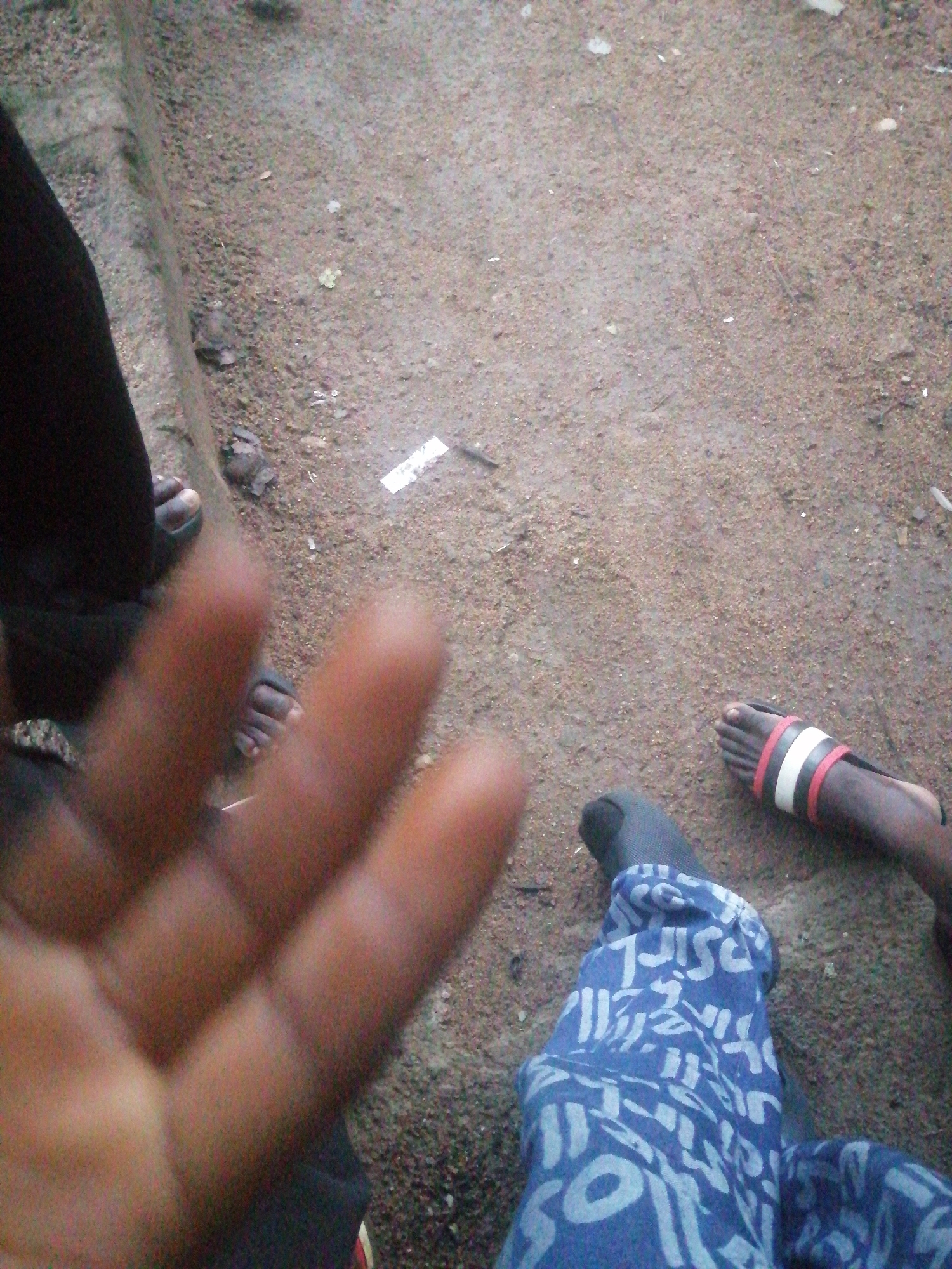 An extreme close-up, shaky, eye-level medium shot shows a hand and feet against a ground made of fine, gritty particles, possibly dirt or sand. The hand, in the foreground, is out of focus and appears to be of a person of color. It is positioned with fingers slightly splayed. Behind the hand, two pairs of feet are visible. The feet on the left are wearing dark sandals. The feet on the right are wearing sandals with red and white straps, and one foot is covered by a blue garment with a white abstract pattern. The background is an out-of-focus view of the gritty ground with scattered debris, suggesting an outdoor environment.