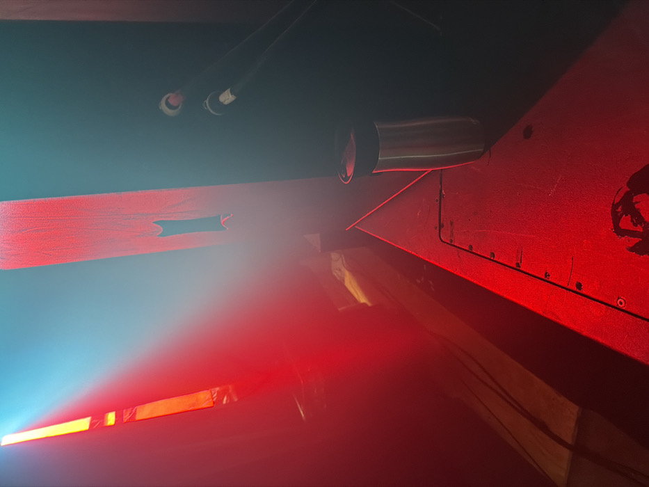 The image depicts an indoor scene characterized by strong artificial lighting and atmospheric haze. In the foreground, an angled, dark-colored surface, possibly a stage monitor or platform, is illuminated predominantly by red light. This surface exhibits multiple small, metallic fasteners along its diagonal edge. A silver-toned cylindrical travel mug with a black lid stands upright on this surface in the mid-right. A dark, irregular mark is present on the lower center of the surface. To the midground left, a vertical, red-illuminated panel, likely constructed of wood, features a dark, irregular cutout near its center. To the midground right, two black cables, each terminating in a circular connector (one with a white band, one with a red band), are connected to or emerge from a dark vertical surface. A broad beam of red light originates from the left, washing over the foreground and the vertical panel. A distinct, narrower beam of blue-cyan light is visible from the upper left, cutting through visible atmospheric haze. The background contains indistinct objects or structures, heavily obscured by red light and haze. No individuals are present in the frame. The specific location within New York City, United States of America, is not identifiable from the visual information provided; the environment is consistent with a performance venue or studio.
