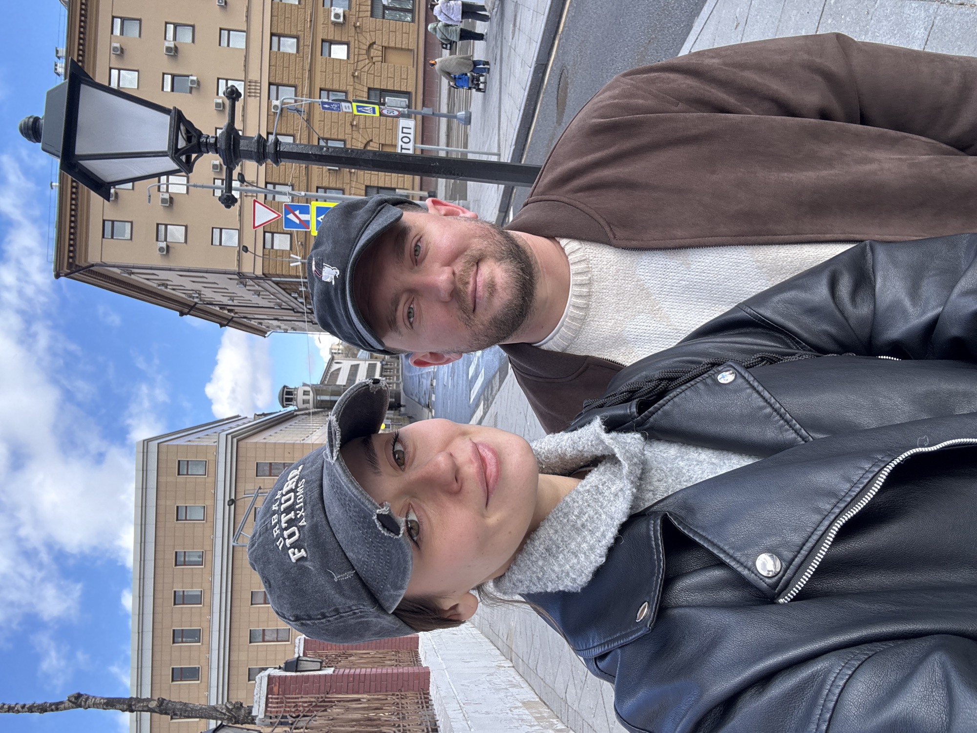 A selfie shot from the perspective of a person wearing a black leather jacket, with a second person standing behind them wearing a brown jacket. Both individuals are wearing caps. The person in the black jacket is wearing a grey cap with text, and the person in the brown jacket is wearing a darker cap with a small logo.
The background shows a streetscape in Moscow, Russia, based on the Cyrillic street sign 'ТОП', and the architecture. A lamppost is on the left. People can be seen walking in the background on the other side of the street. The sky is partly cloudy. The photo is taken from street level.