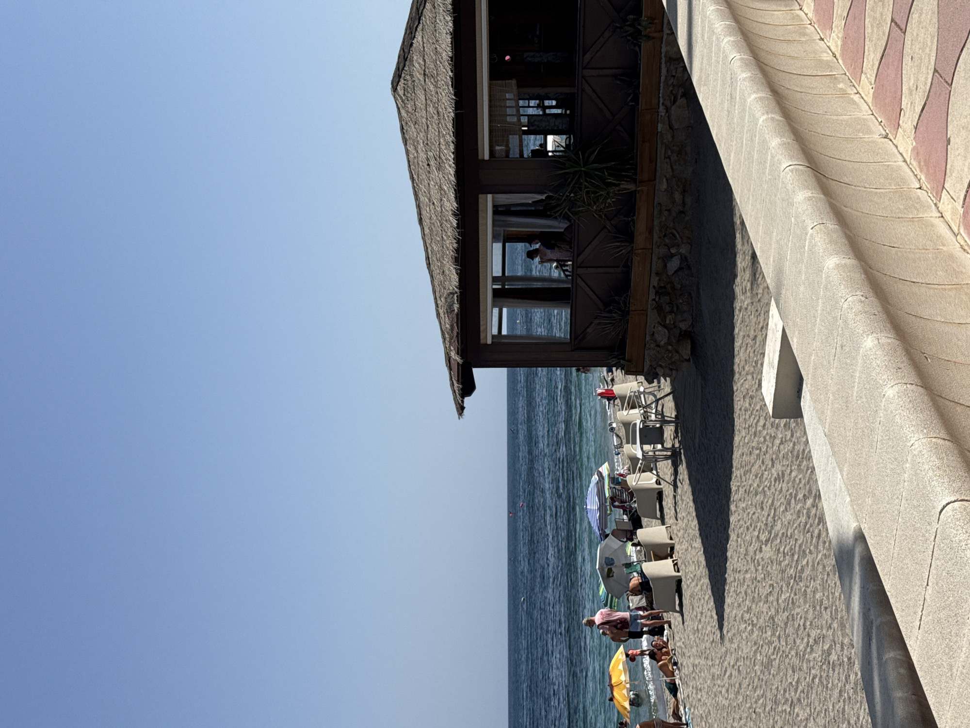 The image depicts a sunny day at a beach in Aguadulce, Spain. In the foreground, a light-colored concrete wall with a curved design runs horizontally across the bottom of the frame, bordering a patterned tiled walkway. Behind the wall, a sandy beach is visible, populated by several beachgoers enjoying the weather. Beach umbrellas, some colorful and some white, are erected over lounge chairs and on the sand, providing shade.
A wooden beachside structure, possibly a bar or restaurant, is situated in the mid-right of the image. It features a thatched roof and large windows offering views of the sea. Inside, silhouetted figures can be seen through the windows.
The sea, a deep blue, stretches across the horizon under a clear, pale blue sky. Small buoys or markers are visible in the water. The overall atmosphere is one of a typical summer day at a Mediterranean beach.