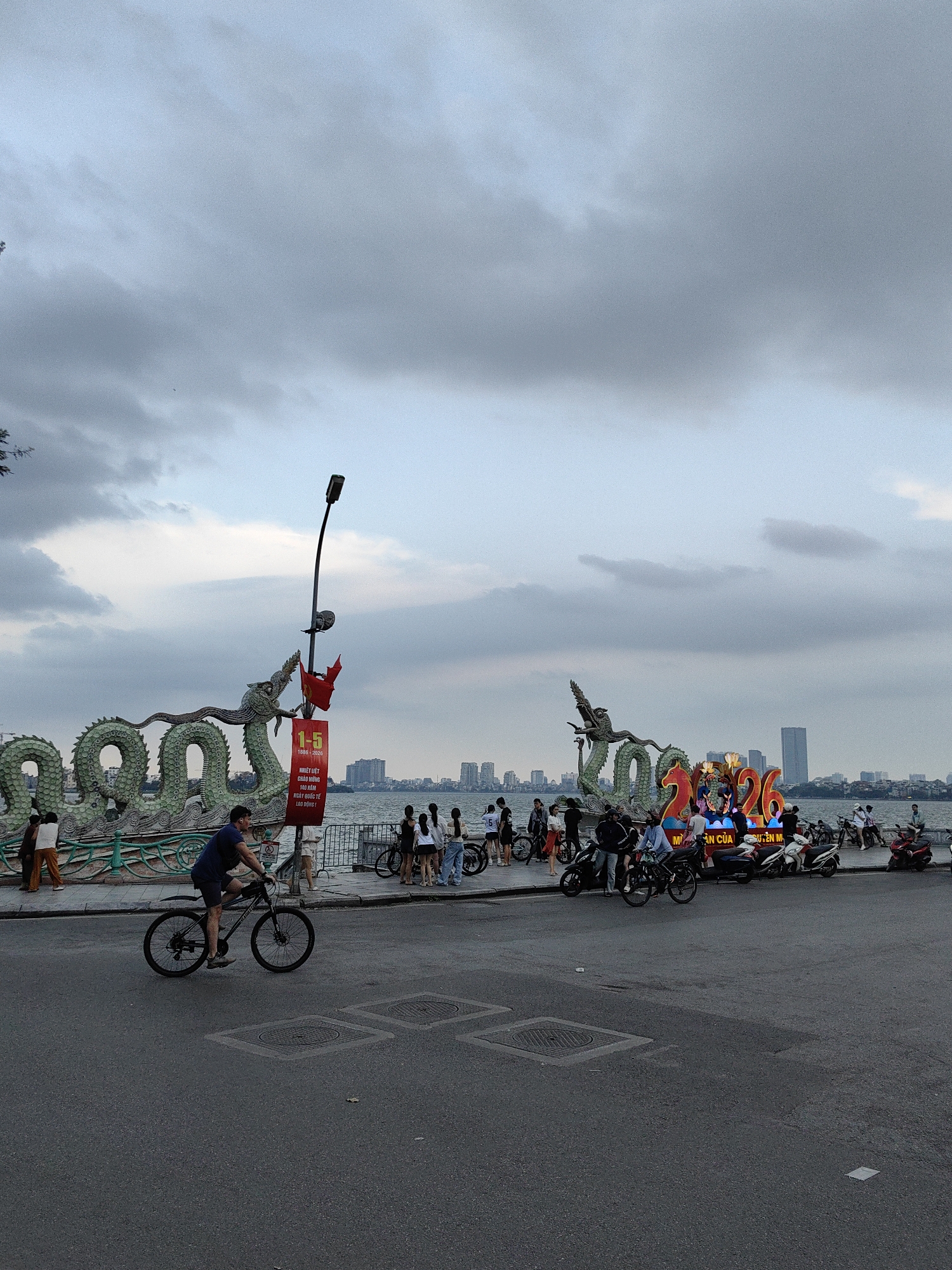 The photograph captures a bustling scene on a city street in Hà Nội, Vietnam, near a body of water that is presumably a lake. In the foreground, a cyclist wearing a dark shirt and shorts is pedaling along the road. The street is paved and shows some manhole covers. On the sidewalk adjacent to the street, a group of people are gathered, with some appearing to be walking or standing. A variety of motorbikes and scooters are parked along the edge of the sidewalk.
Decorative elements are present on the waterfront: large serpentine dragon structures line the promenade, flanking a commemorative display with illuminated numerals "2026". Red flags are visible. In the background, a cityscape is outlined against the sky, with various buildings visible. The sky is overcast with a mix of dark and light clouds. The overall mood suggests a typical evening in an urban setting.