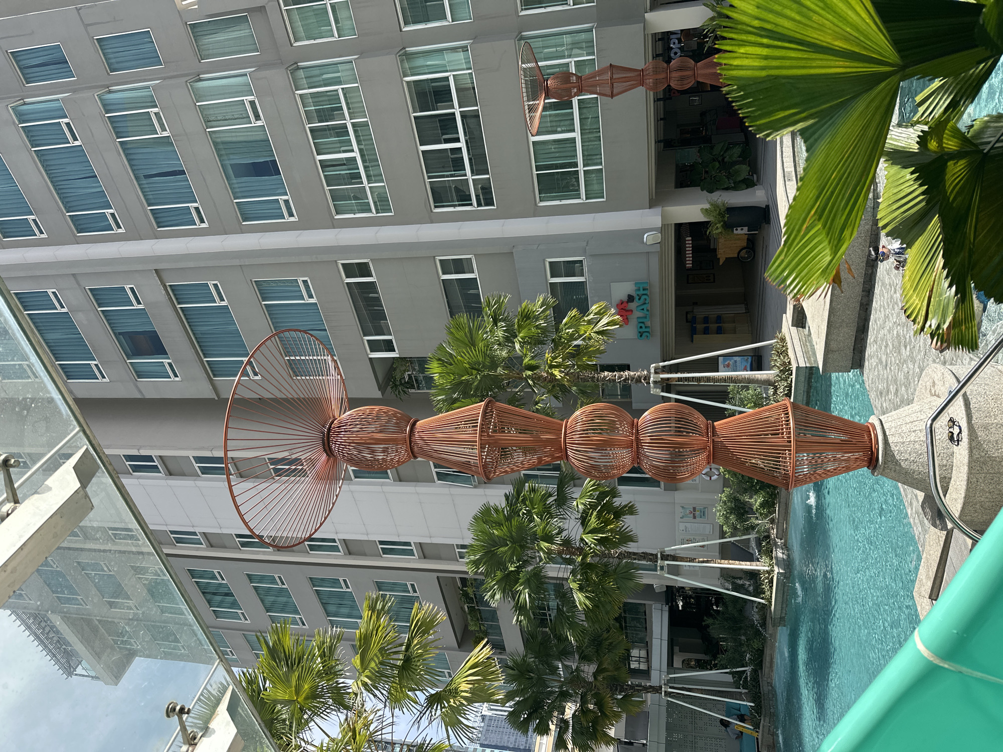 The image depicts a view of a modern building in what is likely Bangkok, Thailand, judging by the architecture and tropical foliage. The building has a gray exterior with multiple rows of windows. A decorative, towering sculpture made of a woven material, possibly bamboo or a similar natural fiber, stands prominently in the foreground and middle ground. The sculpture has a distinctive design, appearing like stacked orbs and cones with a circular fan-like structure at its top. Palm trees and other tropical vegetation are interspersed throughout the scene, adding to the ambiance. Part of a swimming pool is visible at the bottom right, with a turquoise hue. The overall composition suggests a blend of modern architecture with tropical landscaping, common in urban environments within Southeast Asia.