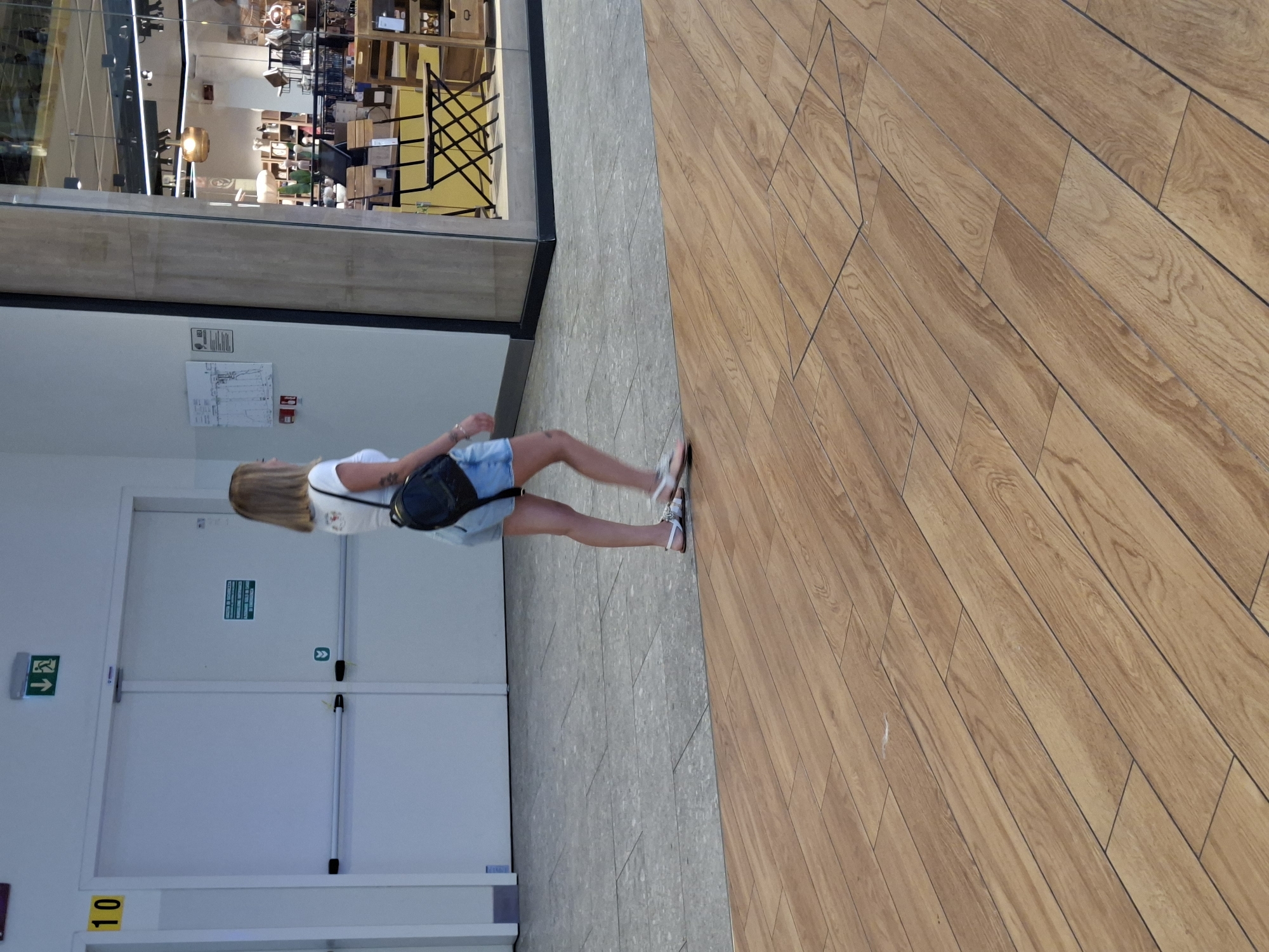 The image shows a person walking in a shopping mall in Arese, Italy. The person is a woman with blonde hair, wearing a white t-shirt and denim shorts, and a black backpack. She is walking on a wooden floor, and in the background, there are doors, a shop window displaying merchandise, and a tiled floor. The scene appears to be indoors, in a commercial setting, and the lighting suggests it could be daytime.