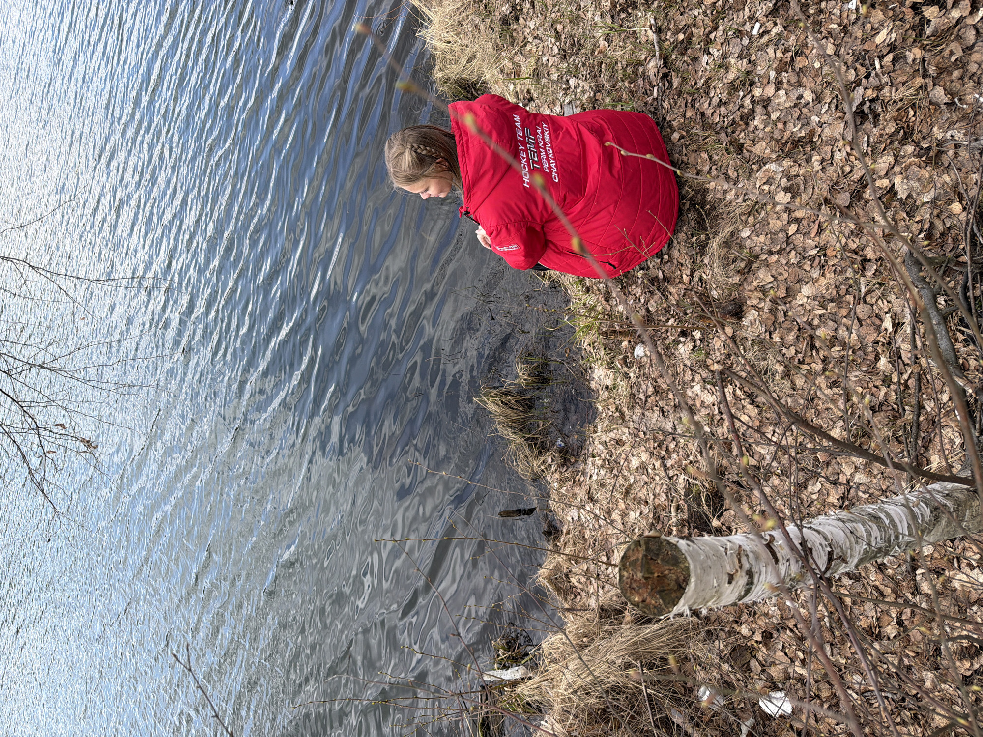 A young girl in a vibrant red padded jacket sits on the grassy bank of a body of water, intently looking down into the ripples. Her blonde hair is styled in a braid. White text is visible on the back of her jacket, reading "HOCKEY TEAM," "TEMPI," "PERM KRAI," and "CHAYKOVSKIY."
The scene is an outdoor, natural setting, likely a lakeside or riverbank in Novyi, Russia. The water, occupying the left side of the image, is a calm blue-grey with numerous small ripples, reflecting bright sunlight that suggests a clear, sunny day, likely late morning or early afternoon. The land on the right is covered with a thick layer of dry, fallen brown leaves and some patches of dry grass. Bare branches and a section of a fallen birch tree with distinctive white, peeling bark are visible in the foreground and on the bank. The overall impression is one of quiet observation by the water's edge during a mild season, possibly early spring or late autumn.