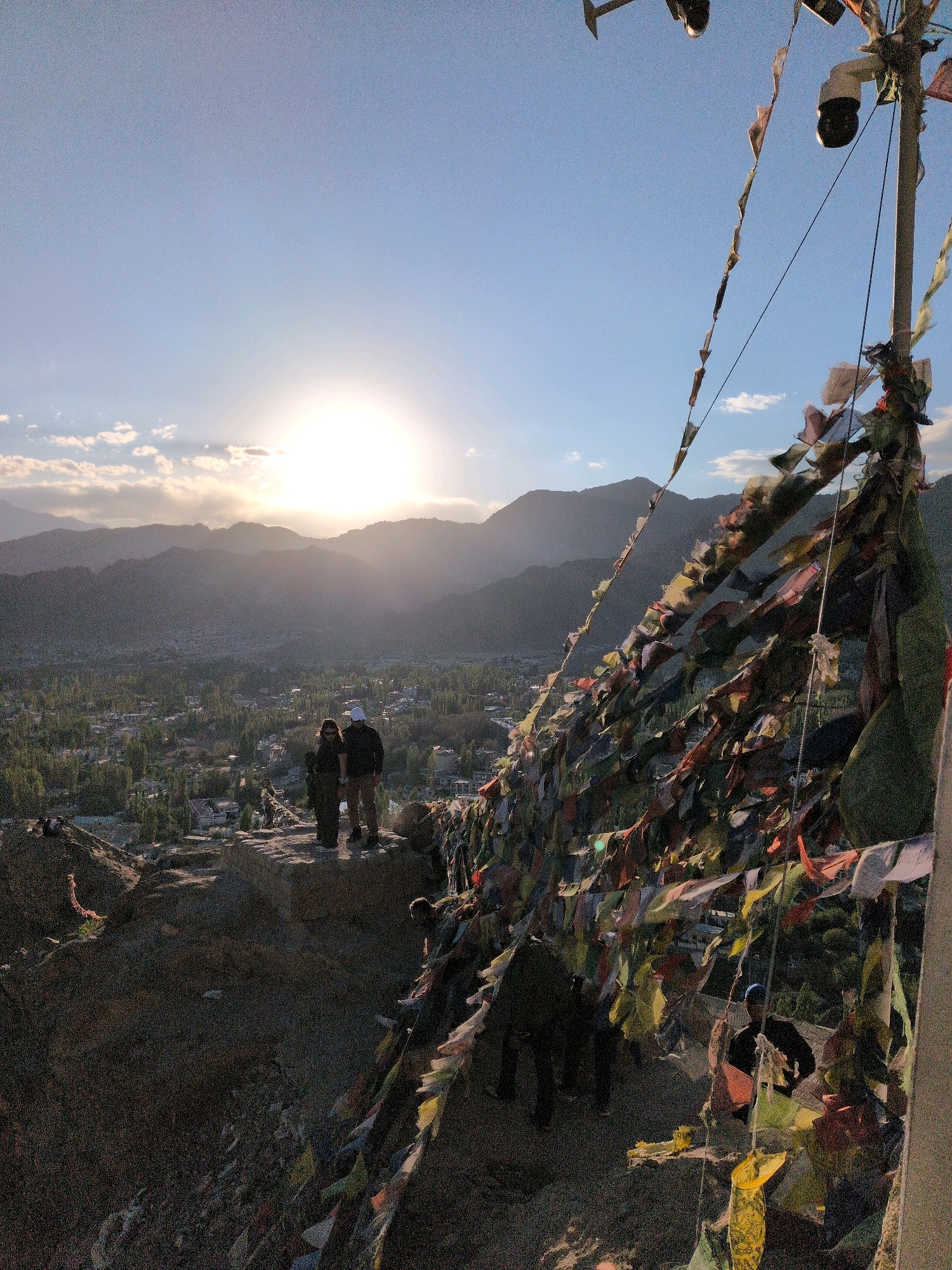 The photograph captures a panoramic view of Leh, India, at sunset. In the foreground, to the right, a tall pole is adorned with numerous colorful prayer flags, creating a vibrant tapestry against the darkening sky. Two individuals stand on a rocky outcrop on the left, silhouetted against the setting sun. They appear to be looking out at the valley below, which is dotted with buildings and lush green trees, indicative of the city of Leh. The sun is low on the horizon, casting a warm, golden light and creating a hazy atmosphere over the distant mountains. The scene evokes a sense of tranquility and the spiritual ambiance of the region.