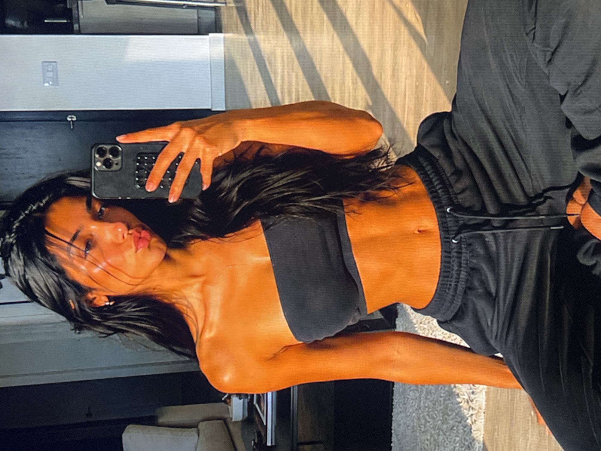 The media file is a vertical, indoor photograph. The main subject is a person, whose face is partially visible as they take a selfie with a smartphone. The person has long, dark hair, and is wearing a black bandeau top and black sweatpants. The lighting suggests it is daytime, with sunlight casting shadows on the floor. The background shows what appears to be a domestic setting, with wooden flooring and a white rug. There are hints of furniture and what looks like a closet or cabinet in the background. The person's skin has a tanned appearance, possibly due to artificial tanning or natural sunlight. The overall mood of the image is casual and personal, typical of a selfie. There is no visible text in the image.