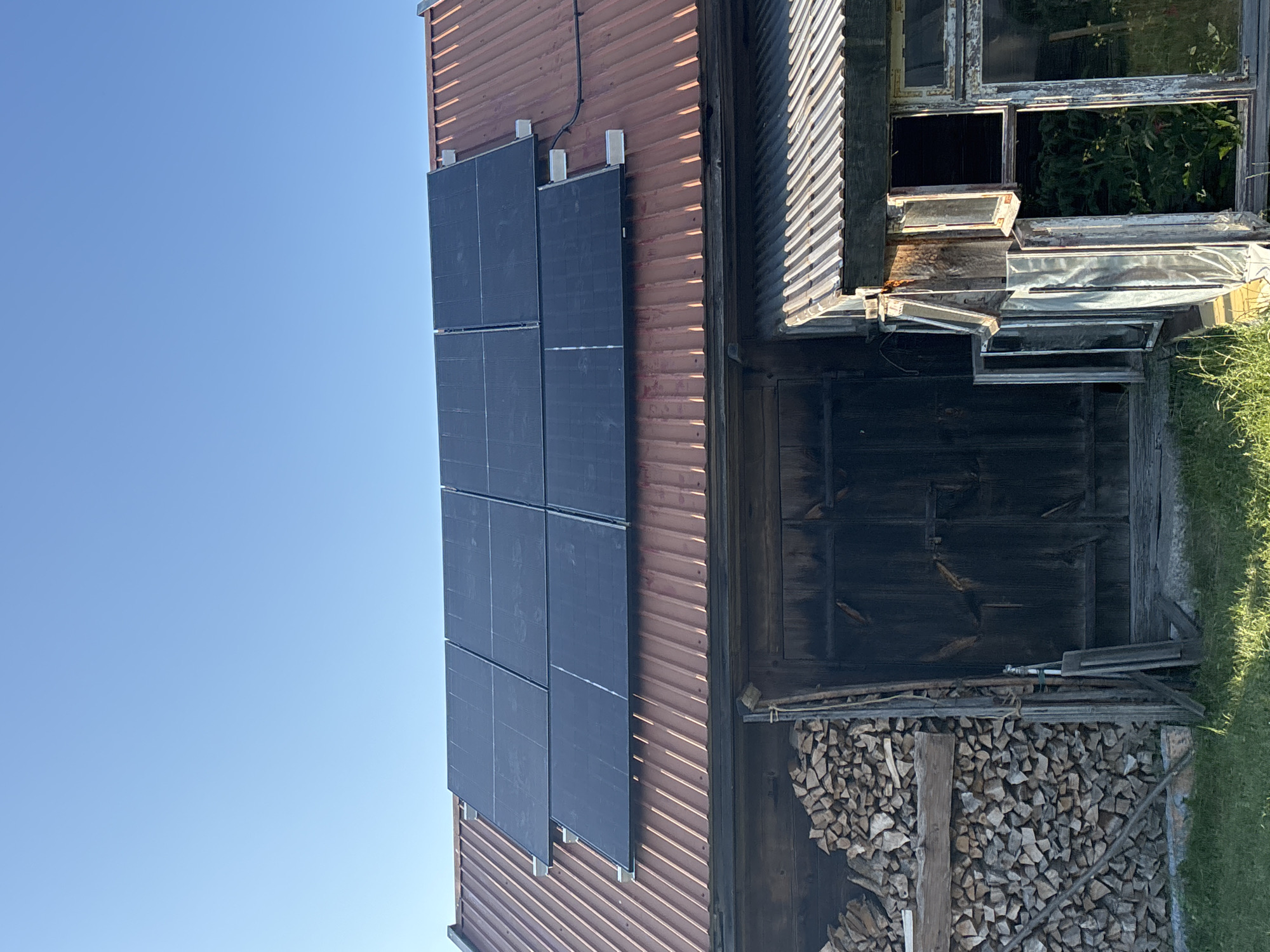 The image shows the exterior of a building with a corrugated metal roof, possibly a barn or shed, located in Janów, Poland. The roof is partially covered with a grid of dark solar panels. Below the roofline, there are wooden walls and large, dark wooden doors. To the left of the doors, a substantial stack of cut firewood is visible. To the right, there is a section of the building with a corrugated metal roof extension and multiple broken or open windows, some of which reveal greenery inside. The sky above is clear and blue. The ground in front of the building is covered with grass.