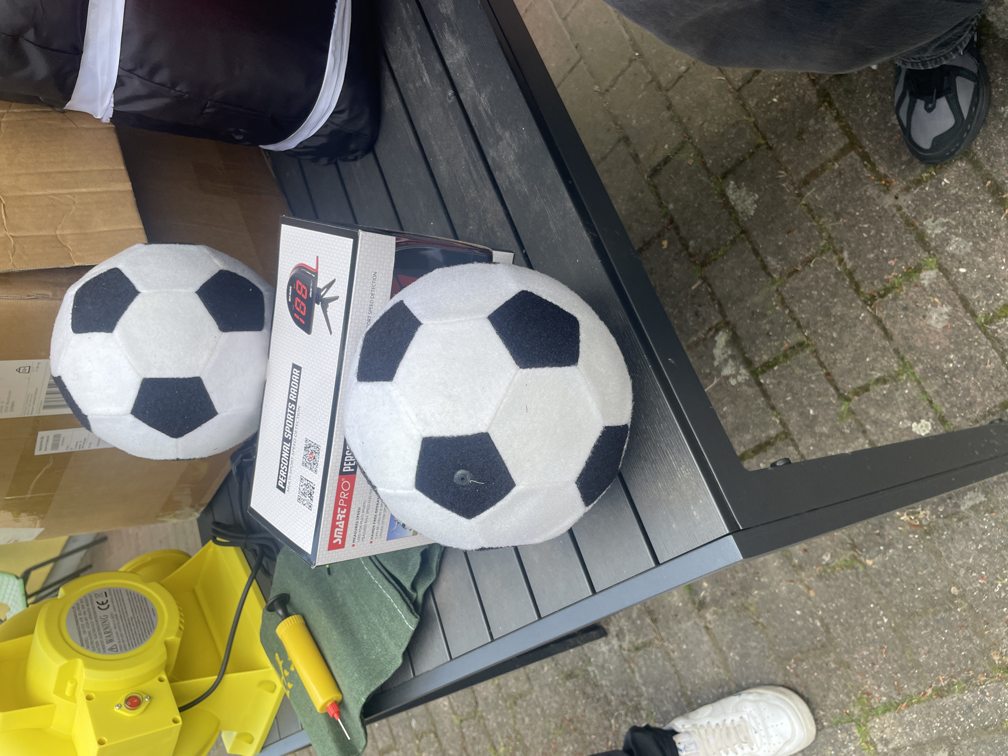 The image shows a black metal table in an outdoor setting, likely a patio or backyard. On the table are two white soccer balls with black patches, the box for a "Smart Pro" personal sports radar, and what appears to be a yellow air pump with a red needle. The text on the box indicates the radar is used for measuring the speed of balls in multiple sports. Part of a black and white inflatable is also visible along with a yellow pump. Some cardboard boxes are sitting nearby. Two people are partially visible. One is wearing black jeans and gray and black sneakers, and is standing to the upper right of the table. The second is wearing black pants and white sneakers and is standing to the lower right of the table. The ground is paved with concrete tiles, and there are signs of grass and weeds growing in the cracks. Based on the weather and background details, the location is Wienhausen, Germany.