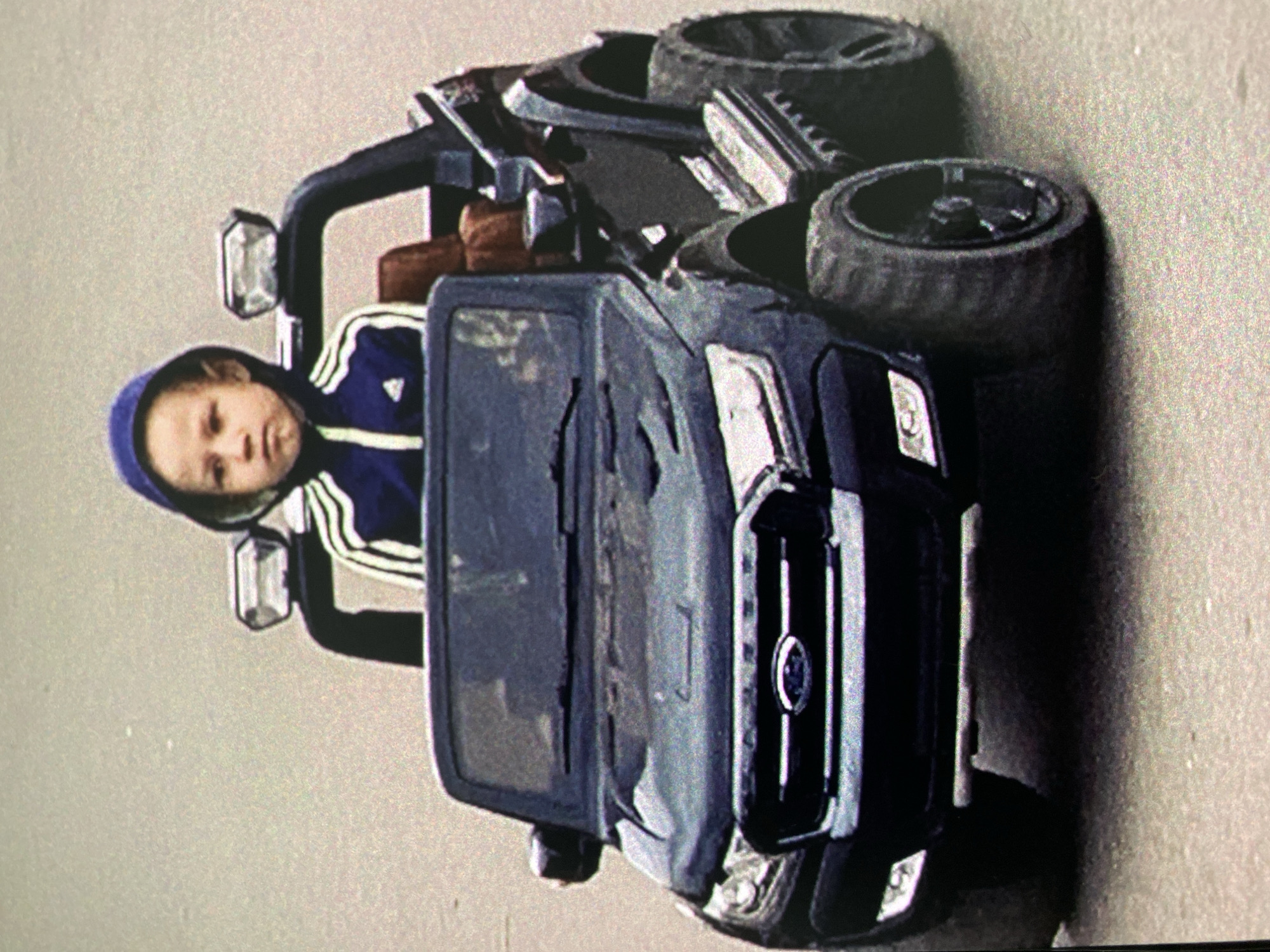 A child wearing a blue Adidas tracksuit sits in a black toy truck. The truck is parked on a gray surface. Two tires rest on the back of the toy truck. The location cannot be determined from the image.