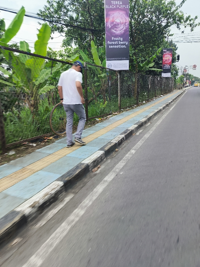 The image shows a man walking on a sidewalk in Padalarang, Indonesia, at coordinates -6.85 latitude and 107.494 longitude. He is wearing a white t-shirt and gray pants, walking towards the right side of the frame. He is walking on a sidewalk with a blue and yellow tiled surface, next to a four-lane asphalt road with a white dividing line. There is a chain-link fence separating the sidewalk from a grassy area with green foliage. Two billboards are visible, one advertising "Terea Black Purple, Frosty forest berry sensation", and the other advertising "Terea Black Purple, Frosty forest berry sensation". A metal bicycle wheel is lying on the ground near the fence. A few vehicles are visible in the distance, driving on the asphalt road. The image is captured from a low angle, with the road and the sidewalk in focus, while the man and the surroundings are slightly out of focus.