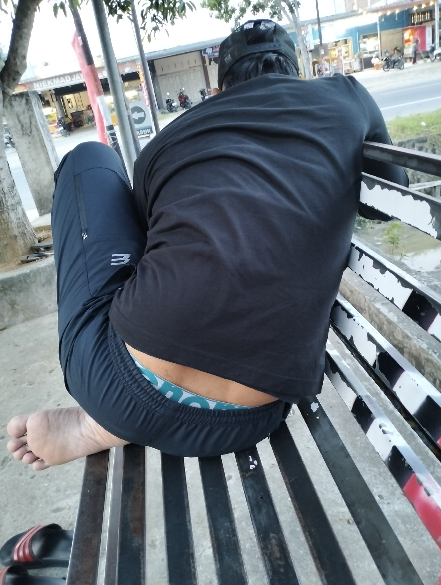 The image depicts a person from behind, seated on a metal bench outdoors, likely in an urban or semi-urban setting. The individual is wearing a black t-shirt and dark athletic pants with a visible light-colored waistband showing. A black baseball cap is worn on their head. Their bare right foot rests on the bench, with a pair of black and red sandals visible on the ground nearby. In the background, there are buildings that appear to be shops or businesses, with signage including the text "NIEKMAD JAY" and "ASUK". There is also a road with some vehicles, including motorcycles, and vegetation. The overall scene suggests a casual outdoor environment, potentially a roadside waiting area or public space.