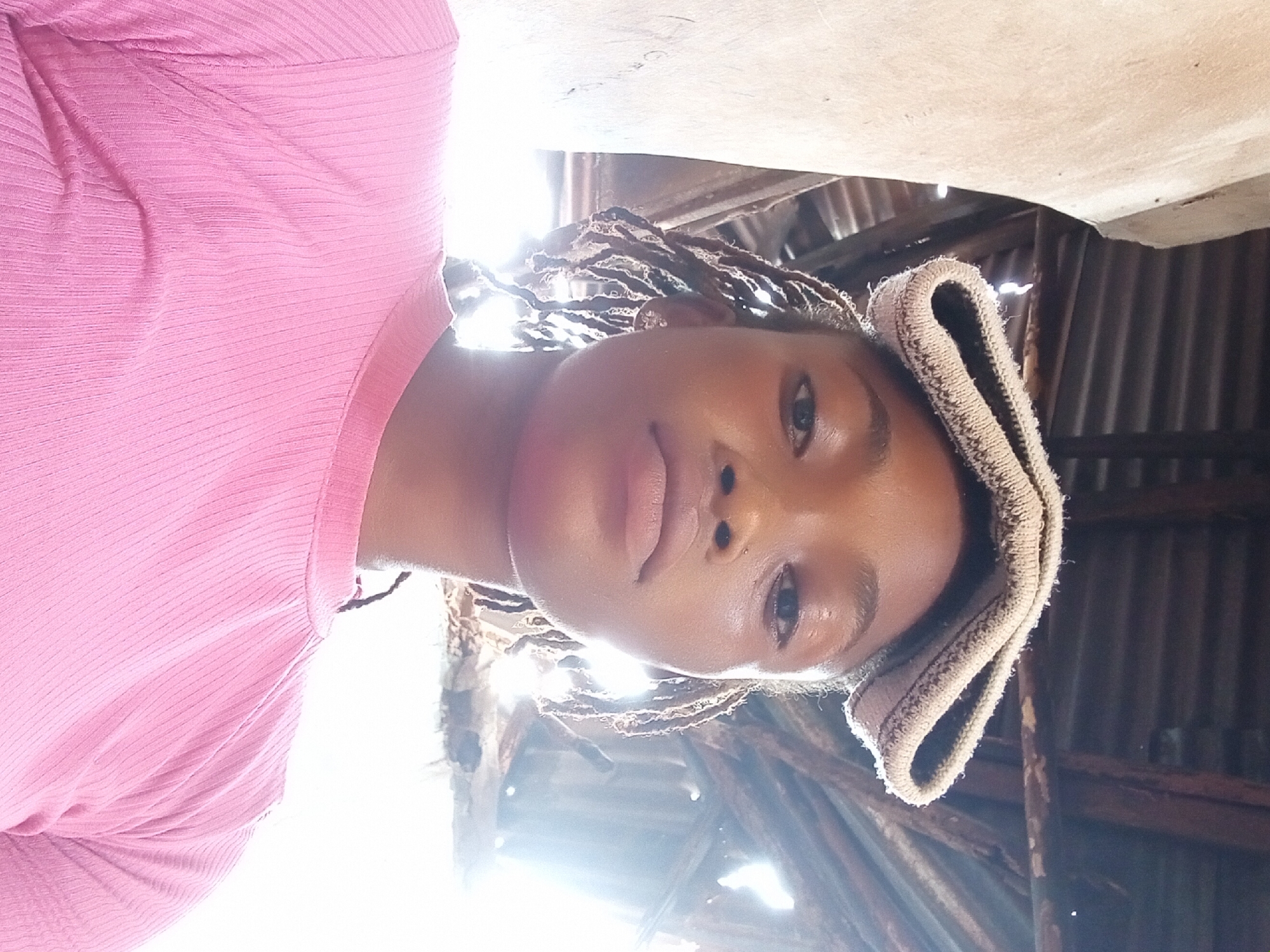 The image is a portrait of a young person, likely female, with dark skin, looking directly at the camera. The person has short, dark braided hair, possibly dreadlocks, which are visible around their face and behind the ears. A light-colored, possibly beige or light brown, textured fabric, resembling a knitted or crocheted head covering or band, is worn on the right side of their head, partially obscuring the right ear.
The subject is wearing a close-fitting pink garment with a ribbed texture, which appears to be a top or dress. The light illuminates their face from the front, highlighting the skin's natural sheen, especially on the forehead, nose, and lips. The expression is neutral and calm, with the eyes directly engaging the viewer.
The background shows what appears to be an indoor or semi-enclosed space, characterized by corrugated metal sheets, likely part of a roof or wall, and wooden structural beams. A strong, bright light source originates from behind the subject and slightly to the left (from the viewer's perspective), suggesting an open window, doorway, or a gap in the structure, indicating daytime. This light creates a bright halo around the subject's head and casts some bright reflections and lens flare, particularly prominent in the upper left and lower central parts of the original image. The overall setting has a rustic or utilitarian feel. There is no visible text in the image.