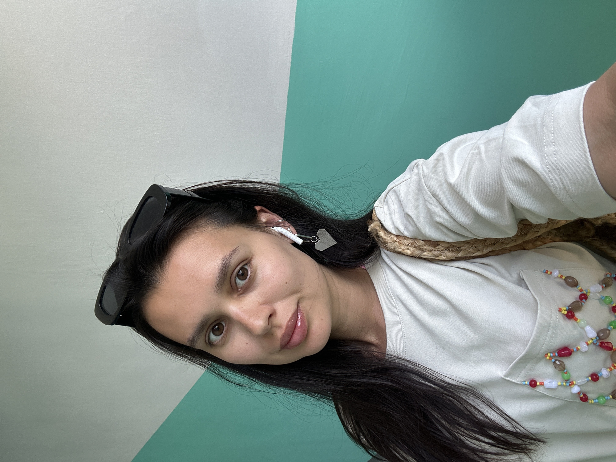 A young woman with long, dark hair is posing for a selfie against a wall painted in two colors, light gray and aqua green. She has sunglasses perched on her head and is wearing a white t-shirt. There's a decorative beaded design on the t-shirt. She has a pair of white earbuds in one ear with a silver heart-shaped pendant hanging down. A woven straw bag is slung over her shoulder. The location is specified as Chaikovsky, Russia, but this detail cannot be verified from the image alone.