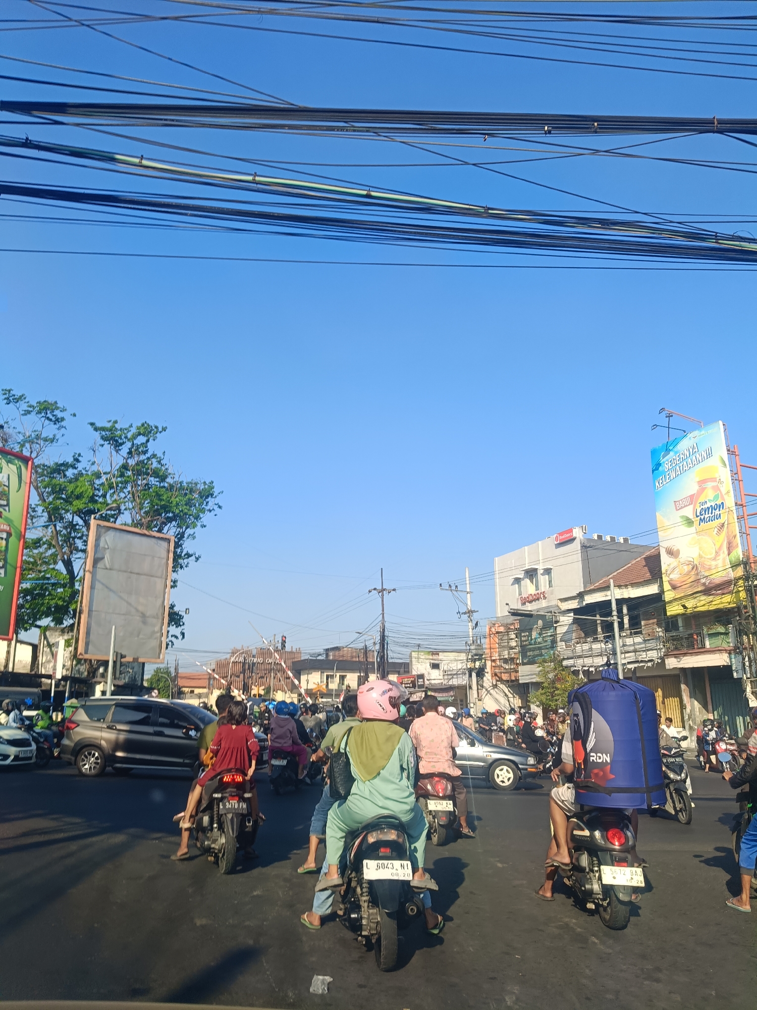 The image shows a street scene in Gedangan, Indonesia, likely during the day given the bright sunlight. A busy street is filled with various vehicles, predominantly motorcycles. Several people are riding on the motorcycles, some with helmets. There are buildings on the sides of the street, including a billboard advertising "Teh Lemon Madu" (Lemon Honey Tea). Overhead, a dense network of power lines stretches across the sky. The sky is clear and blue. The overall impression is of a typical urban street with moderate traffic and commercial activity.