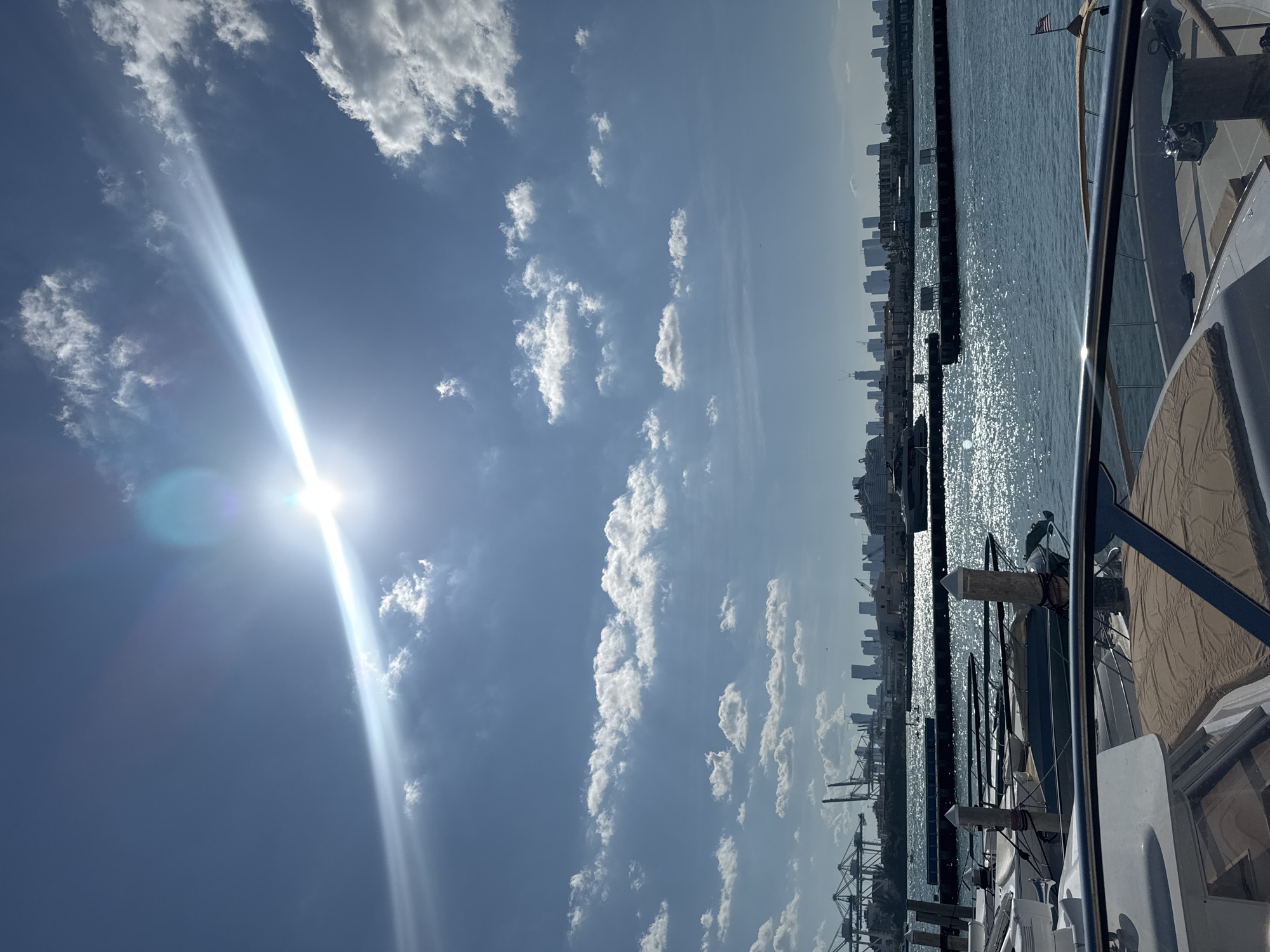 The image shows a view from a boat of the Miami Beach skyline and port in the United States. The sky is mostly blue with scattered white clouds, and the sun is shining brightly. To the right, a glimpse of the boat’s deck and railings can be seen. In the distance, the cityscape is visible along the horizon, and a port with cranes and docked ships occupies the waterfront area. The water reflects sunlight, causing it to shimmer and sparkle.