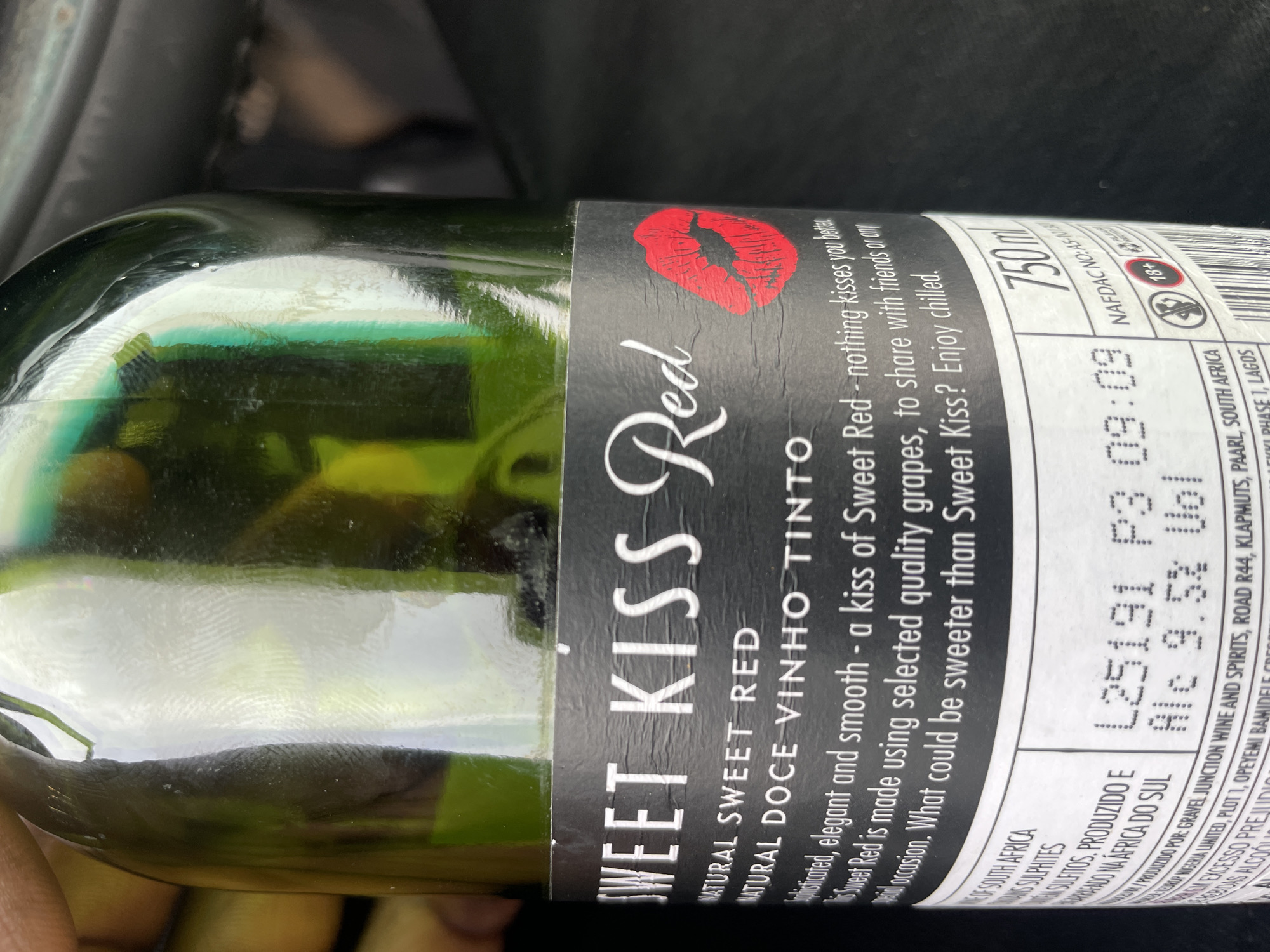 A close-up shot shows a green wine bottle with a black and white label. The label reads "SWEET KISS Red," "NATURAL SWEET RED," and "NATURAL DOCE VINHO TINTO" in large, stylized font. A red lipstick print is visible above the "SWEET KISS" text. Smaller text describes the wine as "sophisticated, elegant and smooth" and suitable for sharing with friends. The label also displays "750ml," a NAFDAC registration number, and an "18+" age restriction symbol. Information at the bottom of the label indicates that the wine is produced in South Africa for Gravel Junction Wine and Spirits, located on Road R44 in Klapmuts, Paarl, South Africa. It is further noted that the wine is imported/distributed by Lamtup Nigeria Limited, Plot 1, Opeyemi Bamidele Crescent, Phase 1, Lagos, Nigeria. A human hand is partially visible at the bottom of the frame. The background is dark and out of focus, suggesting the bottle is inside a vehicle or in a dimly lit area.