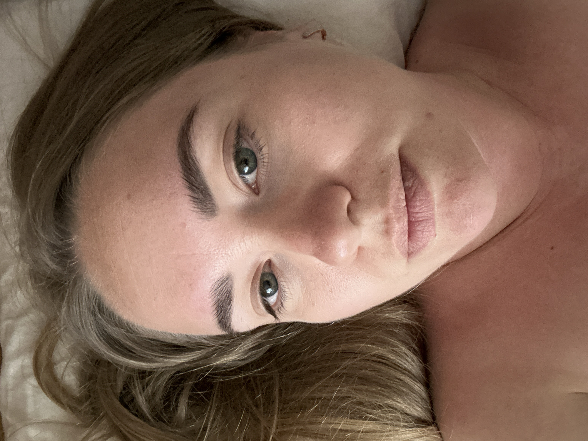 The image captures a close-up, portrait-oriented view of a woman, appearing to be lying down indoors. Her head is tilted, and she looks directly into the camera. She has light brown or blonde hair that frames her face and extends onto what appears to be a white or light-colored pillow or bedding.
Her facial features are prominent, with striking green eyes that hold a calm, direct gaze. Her eyebrows are dark and well-defined. She has fair skin, exhibiting natural texture and a few subtle freckles on her forehead and cheeks. Her lips are slightly parted, hinting at a very subtle, gentle smile. The upper part of her bare shoulder and chest is visible on the right side of the frame, showing natural skin tone with a slight redness.
The lighting in the image is soft and even, suggesting an indoor setting, possibly a bedroom, creating a relaxed and intimate atmosphere. No specific time of day or weather conditions can be determined. There are no visible objects other than the woman and her immediate surroundings. No text is visible within the image. While the provided context is Chaikovsky, Russia, the image itself does not contain any visual cues to confirm this location.