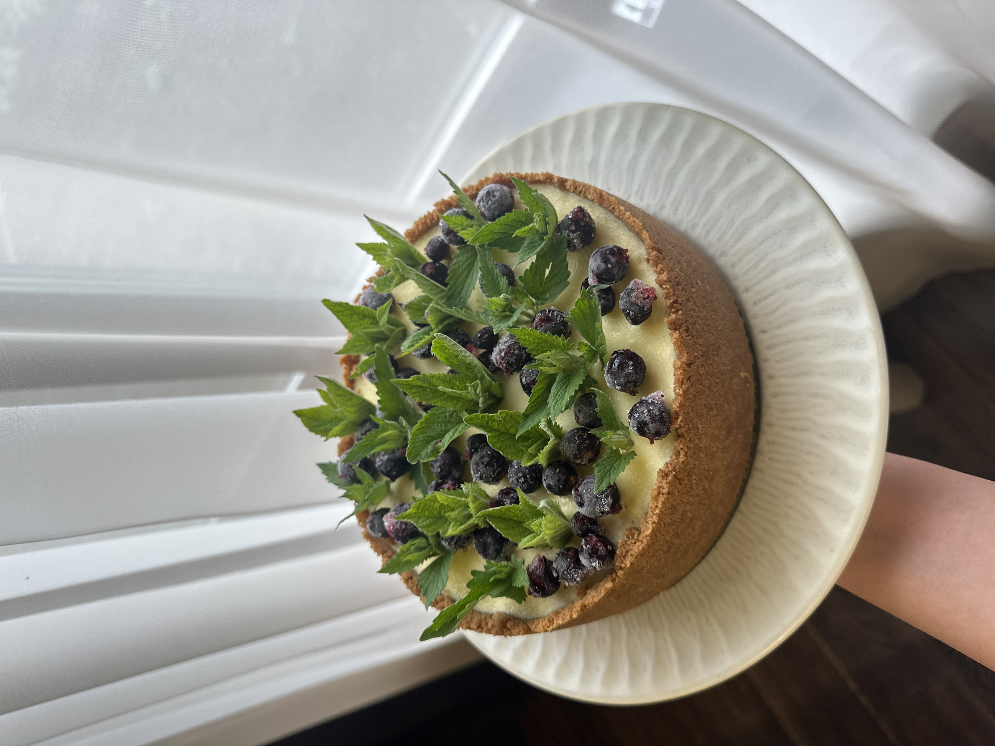 The image displays a round dessert, likely a cheesecake or tart, centered on a light cream-colored ceramic plate. The dessert has a thick, golden-brown crumb crust forming both its base and sides. The filling is a smooth, pale yellow or cream color. The top is generously garnished with dark berries, primarily blackcurrants, many of which appear frosted or lightly dusted with ice crystals, suggesting they are frozen. Interspersed among the berries are numerous fresh, vibrant green mint leaves of varying sizes, arranged for decorative effect. The plate is held by a person's forearm and hand, visible in the lower left foreground, exhibiting fair skin. The background features a large window obscured by vertical white sheer curtains or blinds, creating soft folds and diffusing natural light into the scene. A dark wooden surface, possibly a floor, is partially visible in the lower-left corner. The scene is located in an indoor setting in the city of Chaikovsky, Russia.
