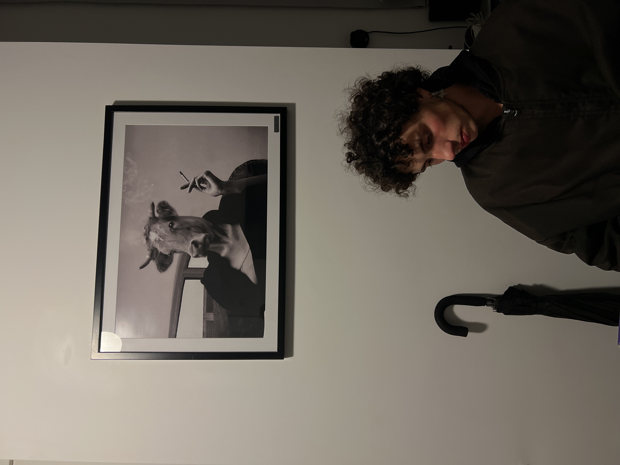 The image depicts an indoor scene against a light grey wall. Centered in the upper half of the frame is a black-framed, monochrome photograph. This artwork portrays a figure with a cow's head, complete with horns, mounted on a human torso. The figure wears a dark, off-the-shoulder garment and holds a lit cigarette in its left hand, from which smoke emanates.
In the lower right foreground, a young individual with dark, curly hair is positioned, looking downwards. This person is wearing a dark, possibly olive-green, zippered jacket over a dark hooded top. A small, silver-colored star-shaped earring is visible in their left ear. There is a reddish mark or flushing on their left cheek and upper lip area.
To the left of the person, a black, folded umbrella with a curved handle hangs on a hook attached to the wall. To the right, near the edge of the frame, a black electrical outlet or device with a cord is visible against the wall. The ambient lighting appears dim, casting soft shadows. No specific architectural or environmental details within the image definitively identify the location as Tel-Aviv, Israel.