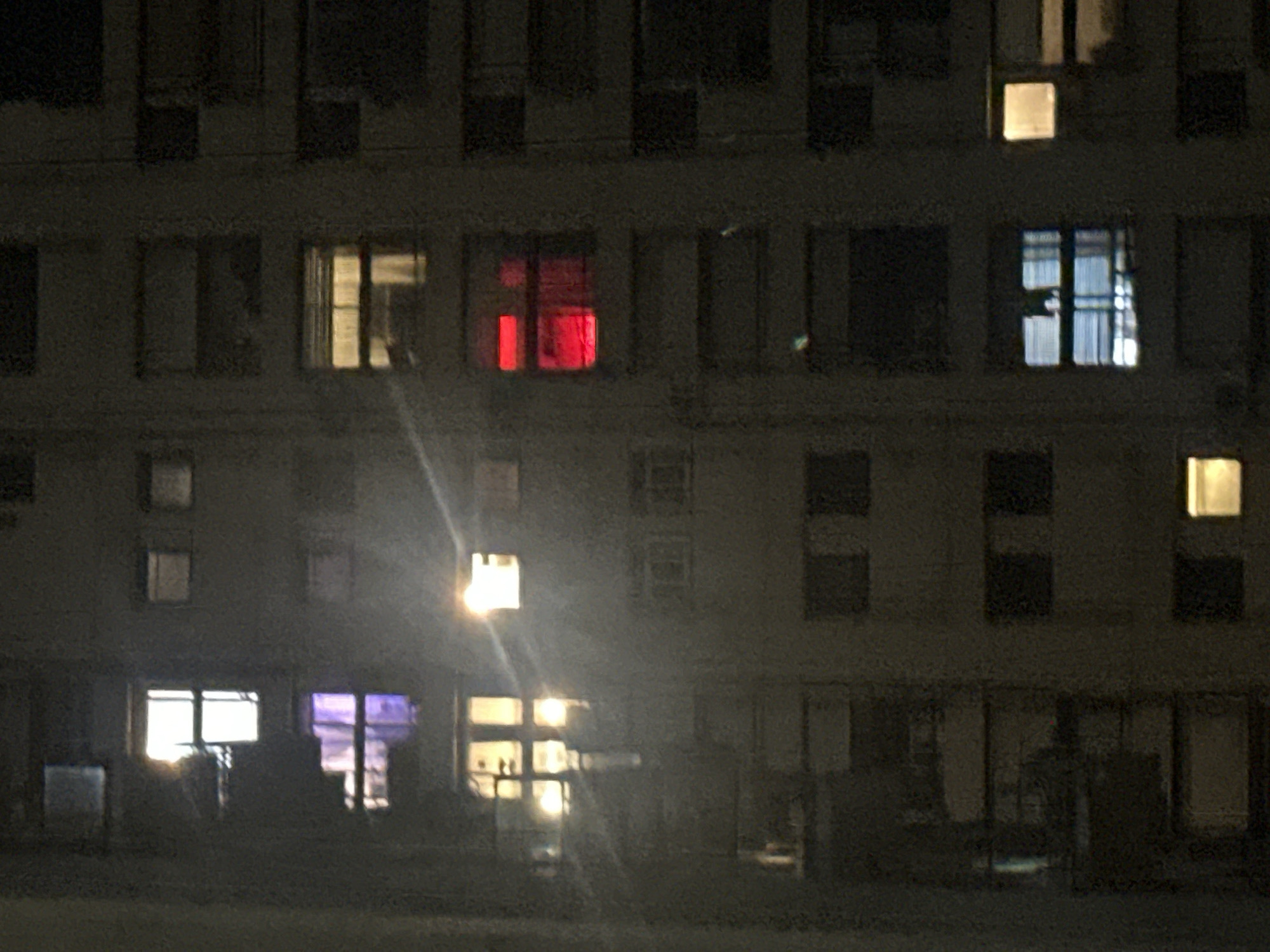 The image depicts a building at night. The building has multiple floors, and several windows are illuminated with different colors: white, yellow, red, and purple. The windows are arranged in a grid-like pattern across the facade. The environment is dark, suggesting it is nighttime. The image appears to be taken from a ground-level perspective. There are no visible people or interactions in the scene. The location is Buenos Aires, Argentina.