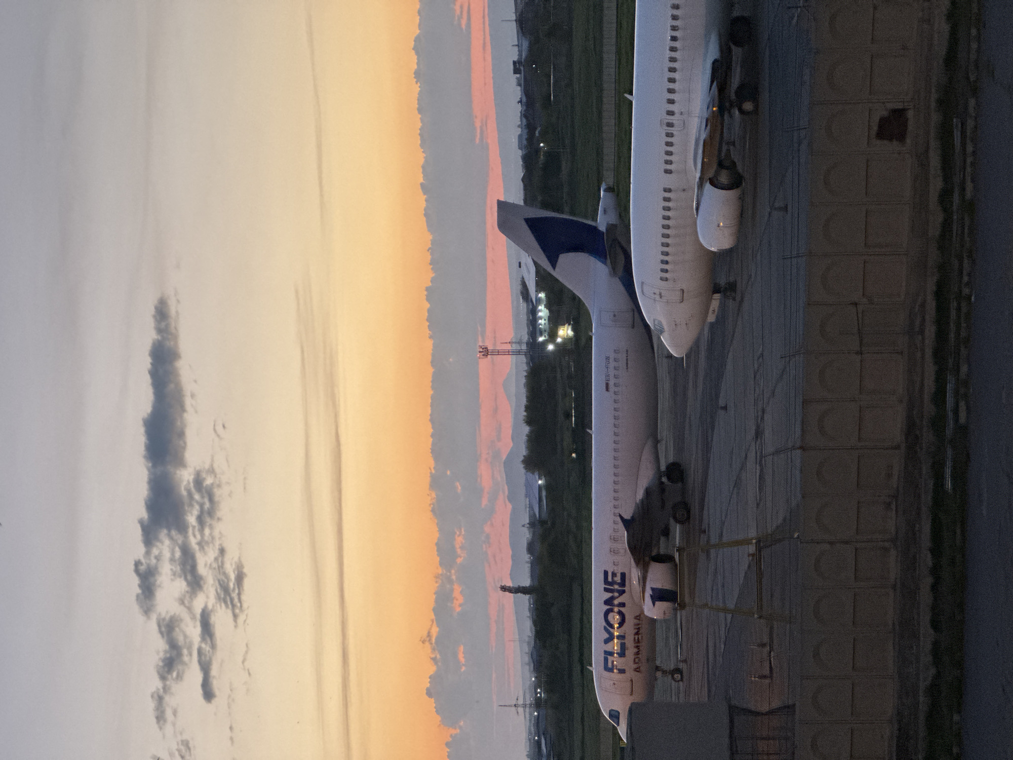 The image depicts an airport tarmac scene during either sunrise or sunset, showcasing the vibrant colors of the sky over Yerevan, Armenia. Two passenger airplanes are parked on the ground. The airplane on the left side of the frame is a white narrow-body jet, clearly bearing the "FLYONE ARMENIA" livery in blue and white letters on its fuselage. Its tail fin is also blue and white. The aircraft type "A320" is partially visible near the front of the plane. To the right, part of a second white airplane, including a section of its fuselage, wing, and engine, is visible, suggesting it is also a parked passenger jet.
The airplanes are situated on a dark concrete or asphalt tarmac, marked with lines and what appear to be large, square concrete slabs. In the background, a low-lying landscape stretches across the horizon, featuring distant buildings, various lights, and a tall communication tower. What might be mountains or hills are subtly visible further in the distance.
The sky is a striking element, filled with the warm, rich colors of dusk or dawn. Hues of orange, pink, and purple blend near the horizon, transitioning to lighter shades of blue and gray higher up. Scattered clouds are present, with one darker, more defined cloud mass visible in the upper left. The scene conveys a sense of calm as the airplanes remain stationary on the tarmac.