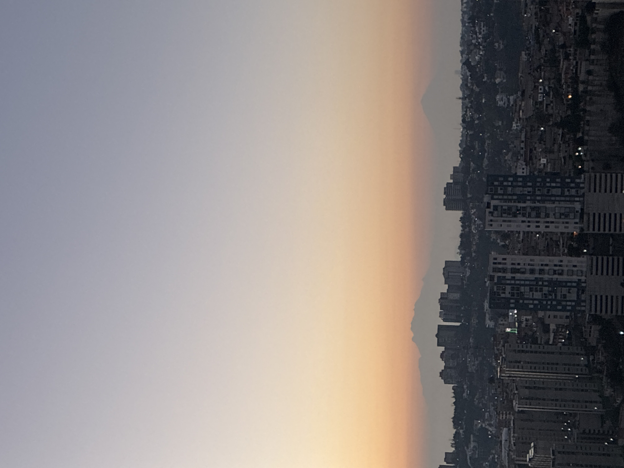 The image shows a cityscape at dusk, possibly Huixquilucan de Degollado, Mexico, with buildings and structures extending towards the right side of the frame. A distant mountain is visible through a hazy atmosphere, situated towards the center of the scene. The sky transitions from a blueish hue on the left to an orange glow closer to the horizon. Buildings appear dark with some hints of artificial lights. The image is oriented in portrait mode, which places the sky above the buildings.