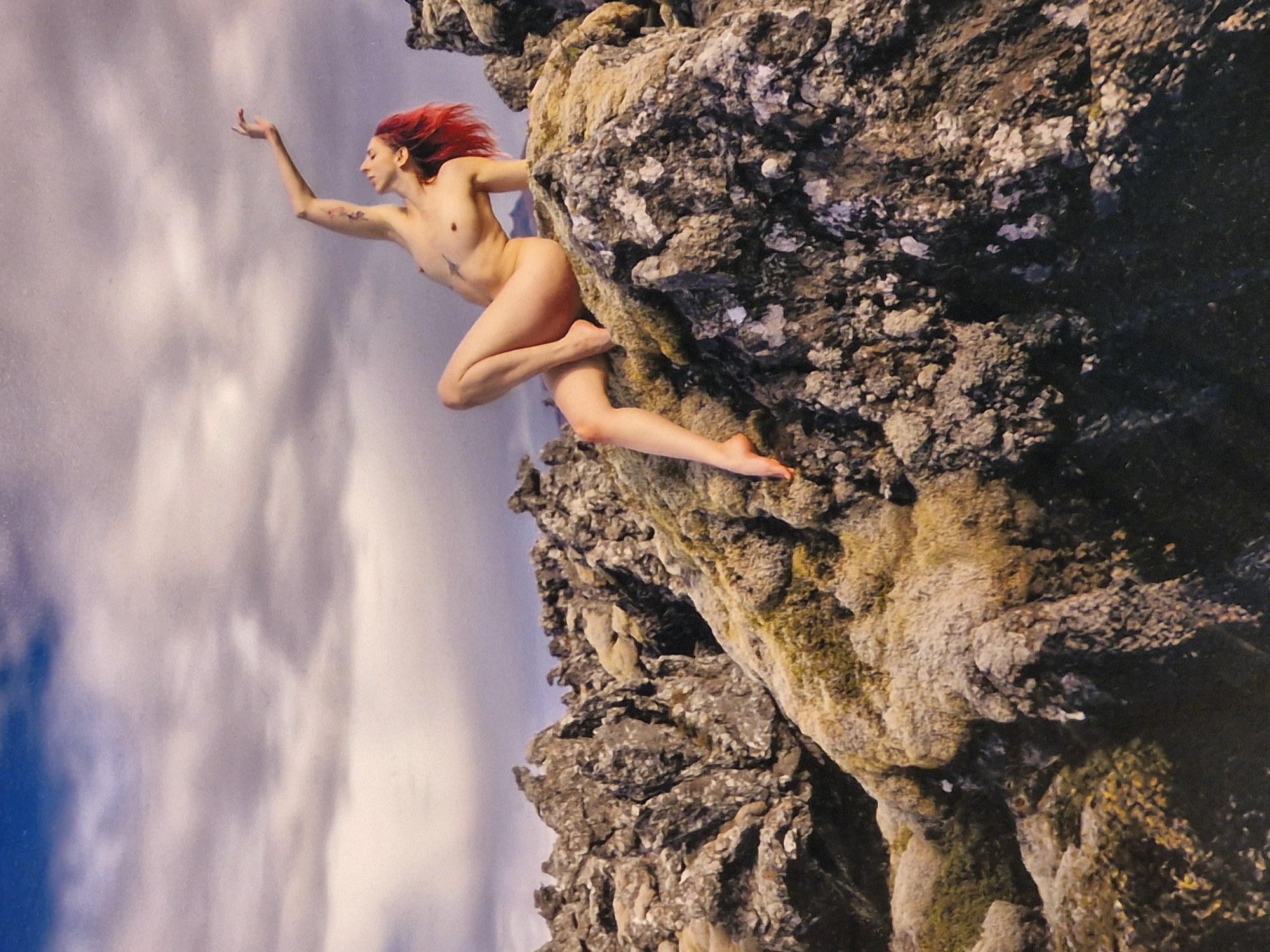 A nude woman with bright red hair is posed on a rocky, moss-covered outcrop. She is leaning back, with her left arm extended upwards and her right arm resting on her bent left leg. She has a tattoo on her left arm and another small tattoo on her chest. The background is a cloudy sky with hints of blue. The rocky terrain suggests a natural, possibly volcanic, landscape. This scene is not located in the city of Bremen, Germany.