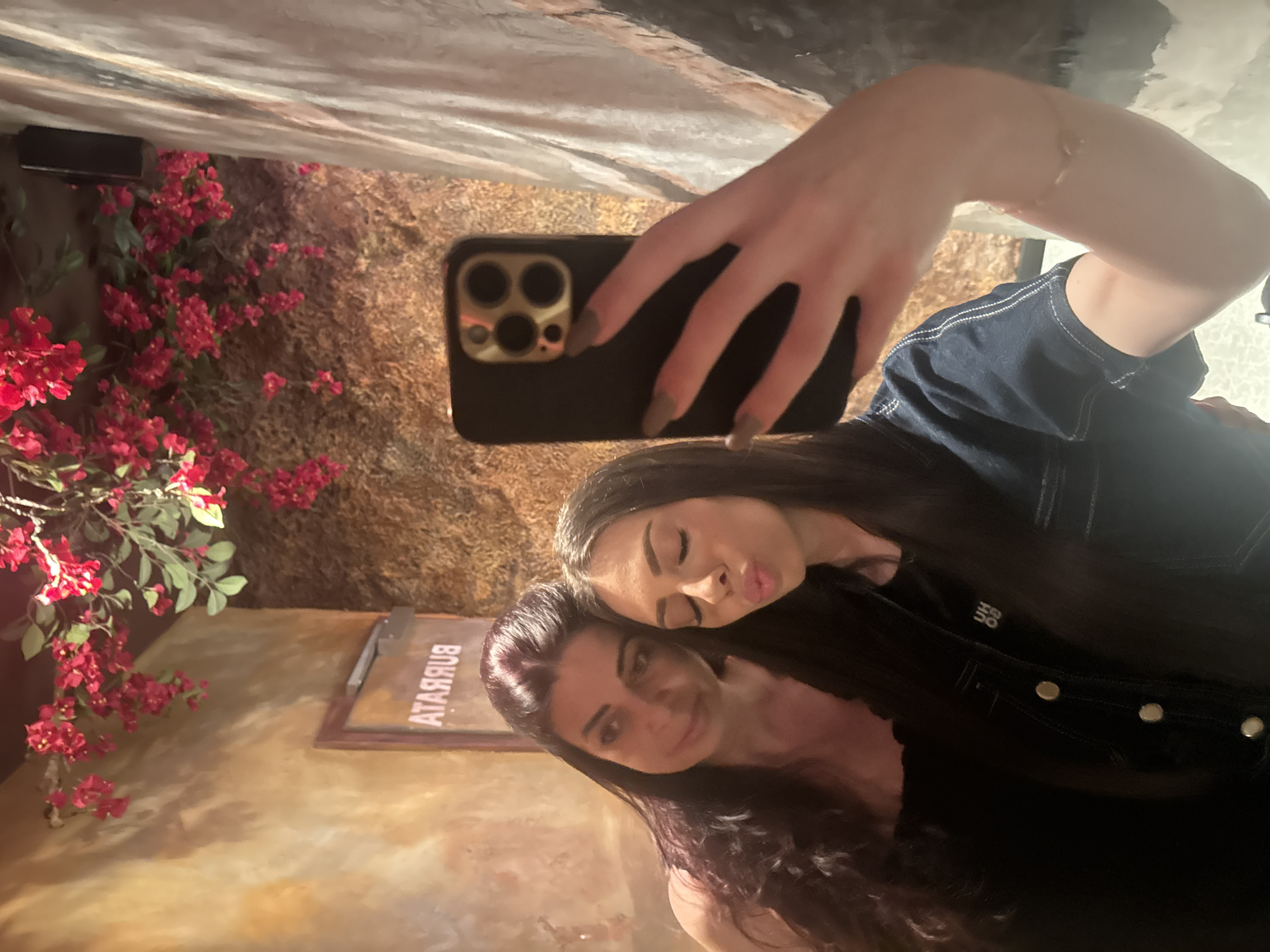 An indoor mirror selfie features two females in a warm-toned, dimly lit establishment. The woman on the left, appearing older, has long dark hair and wears a dark top. She looks towards the right. The woman on the right, appearing younger, has long dark hair, a denim shirt/jacket with a small white pin displaying "UH OO" (or similar text) on her lapel, and light brown painted nails. She holds a black smartphone with gold camera bezels in her right hand, taking the selfie while making a kissy face. A delicate bracelet is visible on her left wrist.
The background consists of textured walls: a warm, ochre-toned wall on the upper left, and a darker, possibly moss-covered or rough-textured wall on the upper right and center-right. Abundant artificial red/pink floral decorations and green foliage hang from the ceiling and walls. A sign, reading "ATARRUB" (reversed, unreversed "BURRATA"), is visible on a door or frame behind the woman on the left. A reflective surface, likely a mirror or polished material, runs vertically along the far right edge of the frame.
There are no visual cues within the image to confirm the location as Varna, Bulgaria.