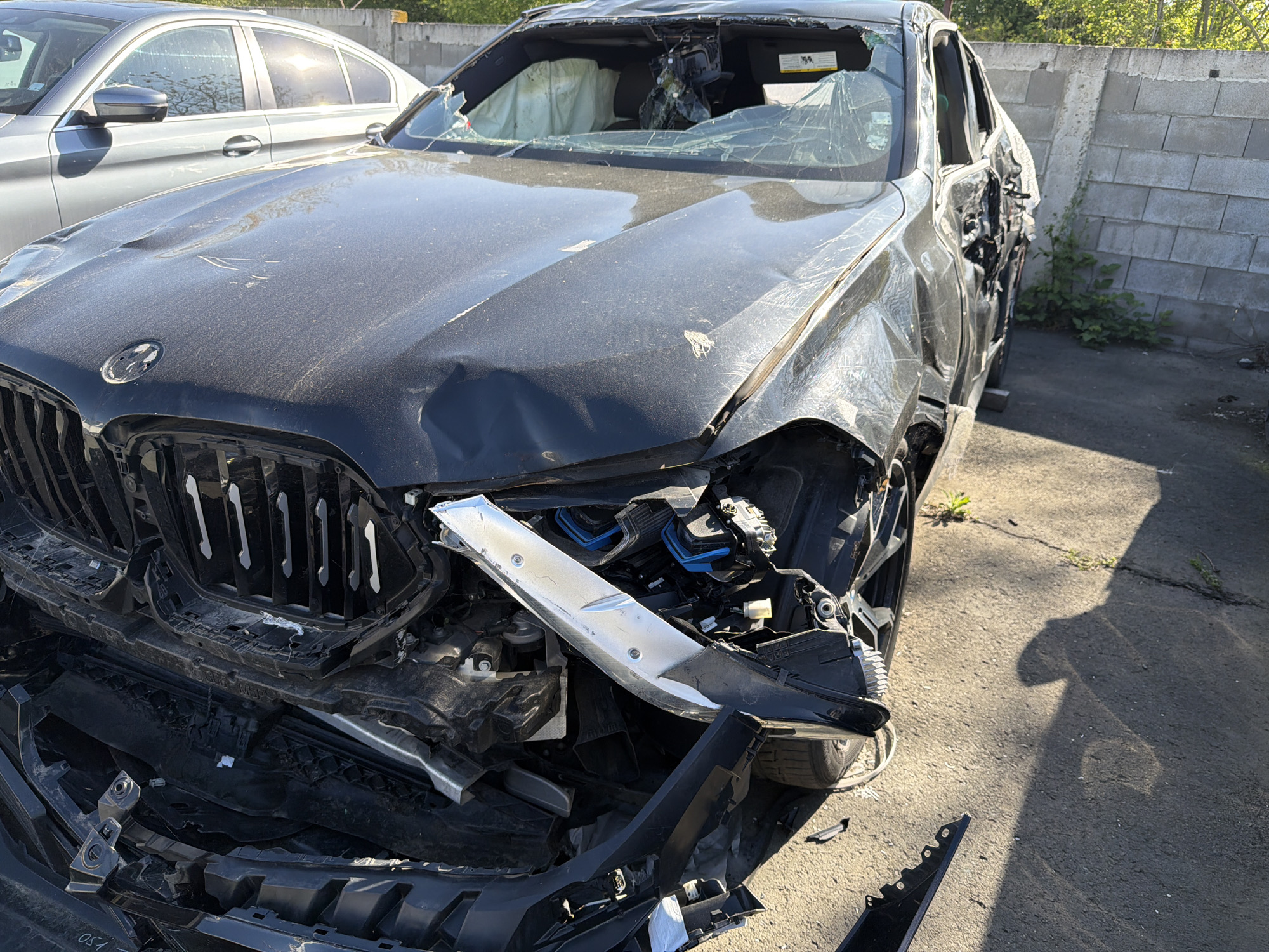 The image shows a black BMW with extensive damage to the front end, including a crushed hood, broken windshield, and missing bumper. The driver's side door is also heavily damaged. The car is located on a paved surface next to a concrete block wall. There is another car parked behind the damaged BMW. The location is in the city of Unknown, Unknown.