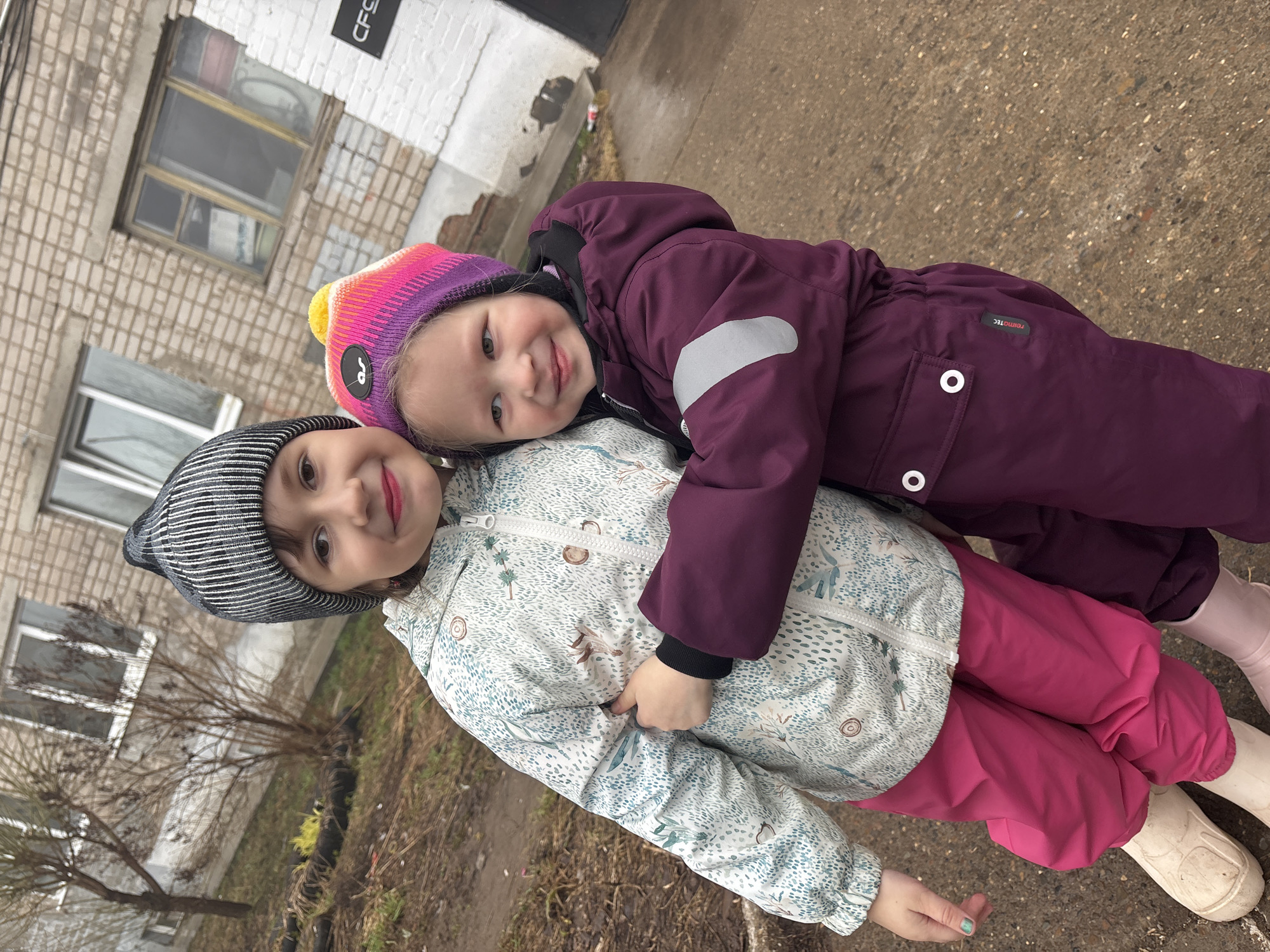 Two young girls are posing for a photo outdoors. One girl is wearing a white jacket with a leaf and branch pattern, pink pants, a black and white striped beanie, and light-colored boots. The other girl is wearing a purple jacket, purple pants, a colorful striped beanie, and light-colored boots. The girl in purple is lying on top of the girl in the white jacket, with her arm around her. They are both smiling at the camera. The background includes a building with brick walls and windows. Based on visual cues such as the architecture, the environment (bare trees, possibly thawing ground), and information available on the building in the background, it is highly probable the photo was taken in the city of Chaikovsky, Russia. The ground appears to be a mix of dirt, grass, and pavement. The weather seems to be cold.