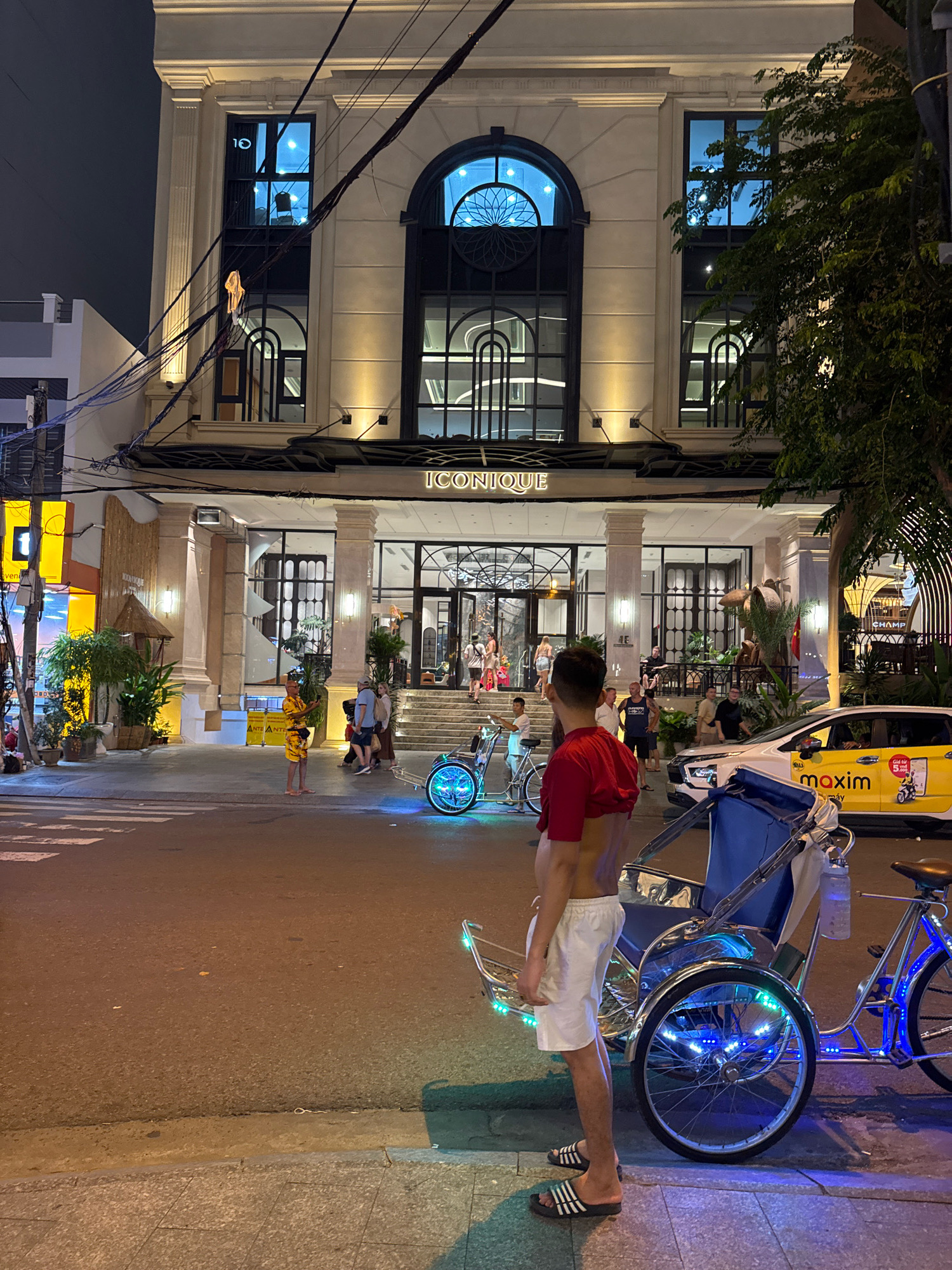 A street scene at night in Nha Trang, Vietnam, is depicted. The main focus is a man in a red t-shirt and white shorts standing next to a cyclopousse, a three-wheeled vehicle for transporting passengers, which is illuminated with blue LED lights. In the background, a modern building with large windows, the "ICONIQUE" sign, and an entrance with stairs is visible. Several people are seen walking on the street and entering the building. A yellow taxi with "maxim" branding is parked on the right side of the street. Trees and streetlights illuminate the scene.